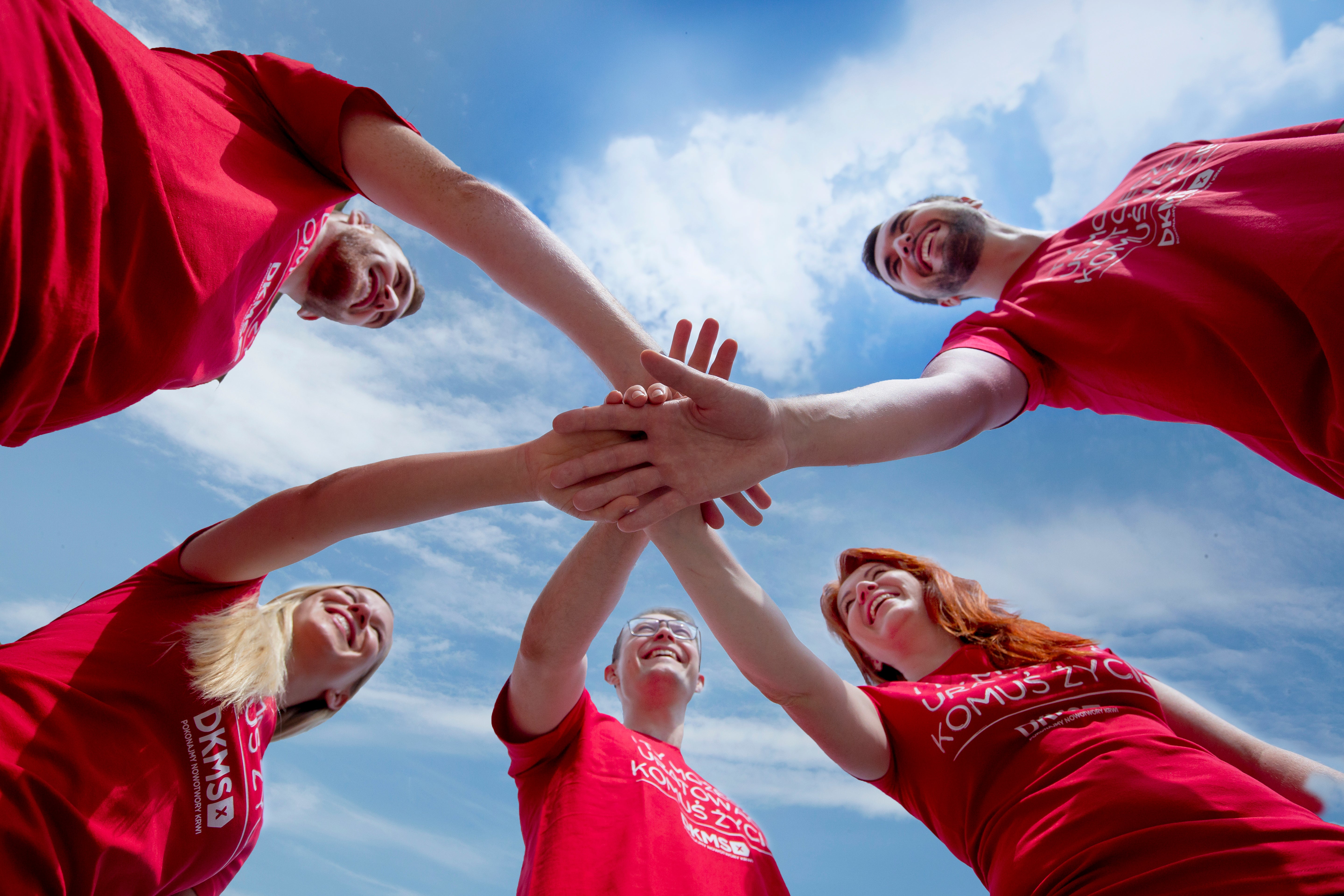 A group of five people in red DKMS shirts putting their hands in the middle together. You can see them from below. The sky above them is blue with some clouds and sunny weather.