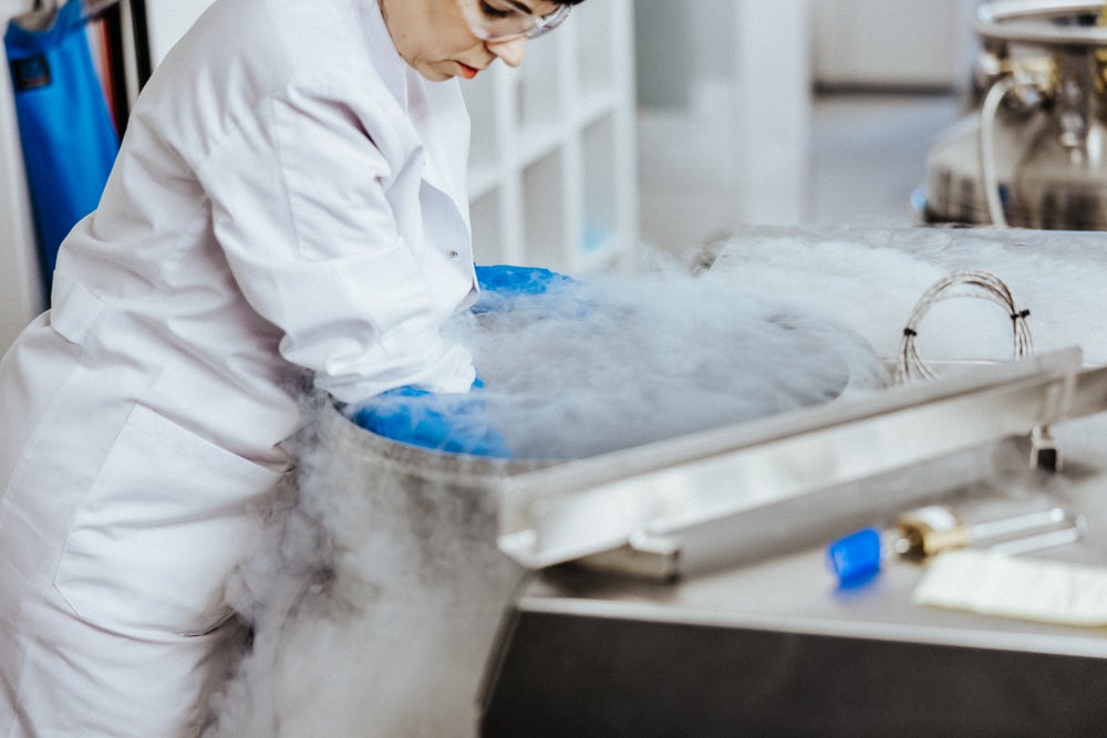 A female scientist from the DKMS Life Science Lab, wearing a white lab coat, safety goggles, and blue cryogenic gloves, is removing a storage rack with stored stem cell preparations (ADCU) out of a storage tank surrounded by vapor.