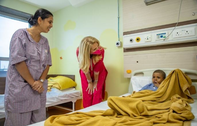 A young boy lies in a hospital bed covered with a yellow blanket at a Sankalp BMT Unit. The CEO Elke Neujahr is standing by the bed and and leaning forward to engage with the boy. She is wearing a bright pink outfit and a pink mask. The other woman, dressed in a hospital gown, appears to be the boy's mother. The hospital room is softly lit and welcoming.