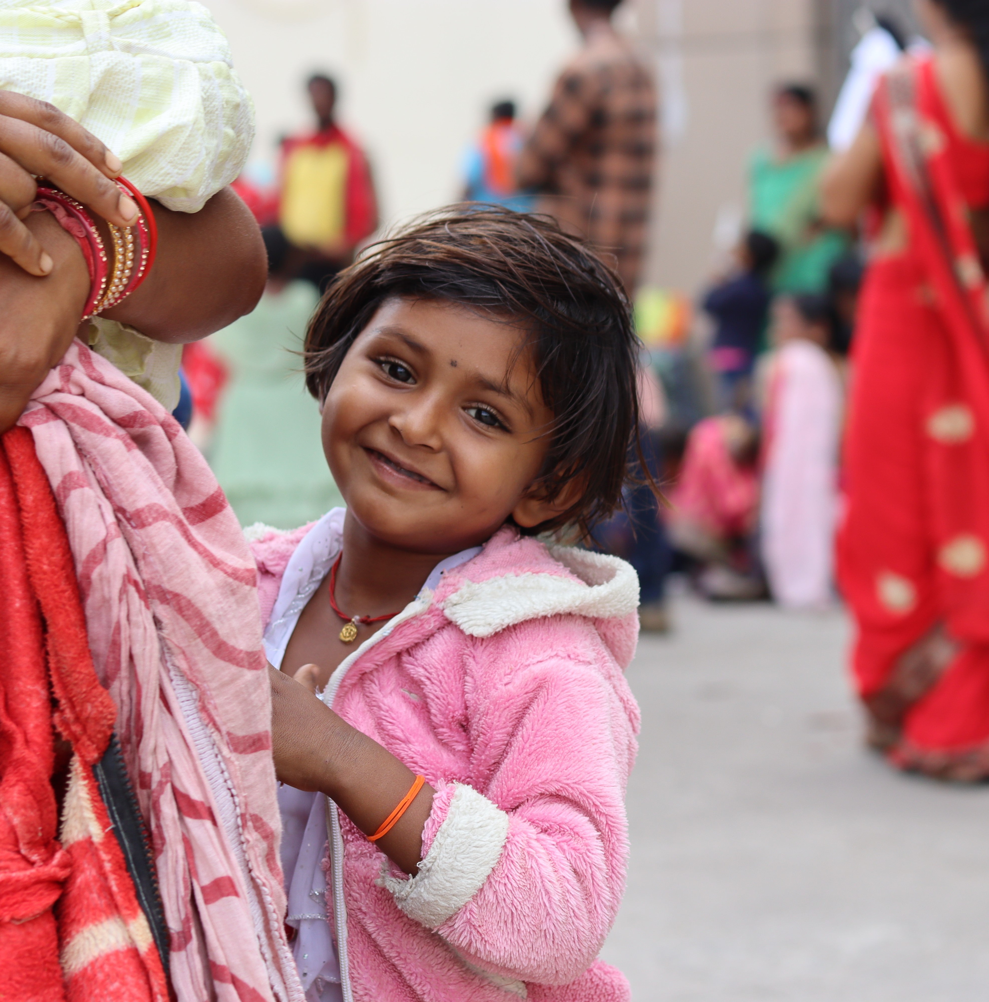 A little Indian girl, who is smiling and wearing a pink hoodie, is standing next to a woman of whom only one arm and the hand are visible. The woman is holding some clothes in her hand. In the background, people in colorful clothing can be seen.