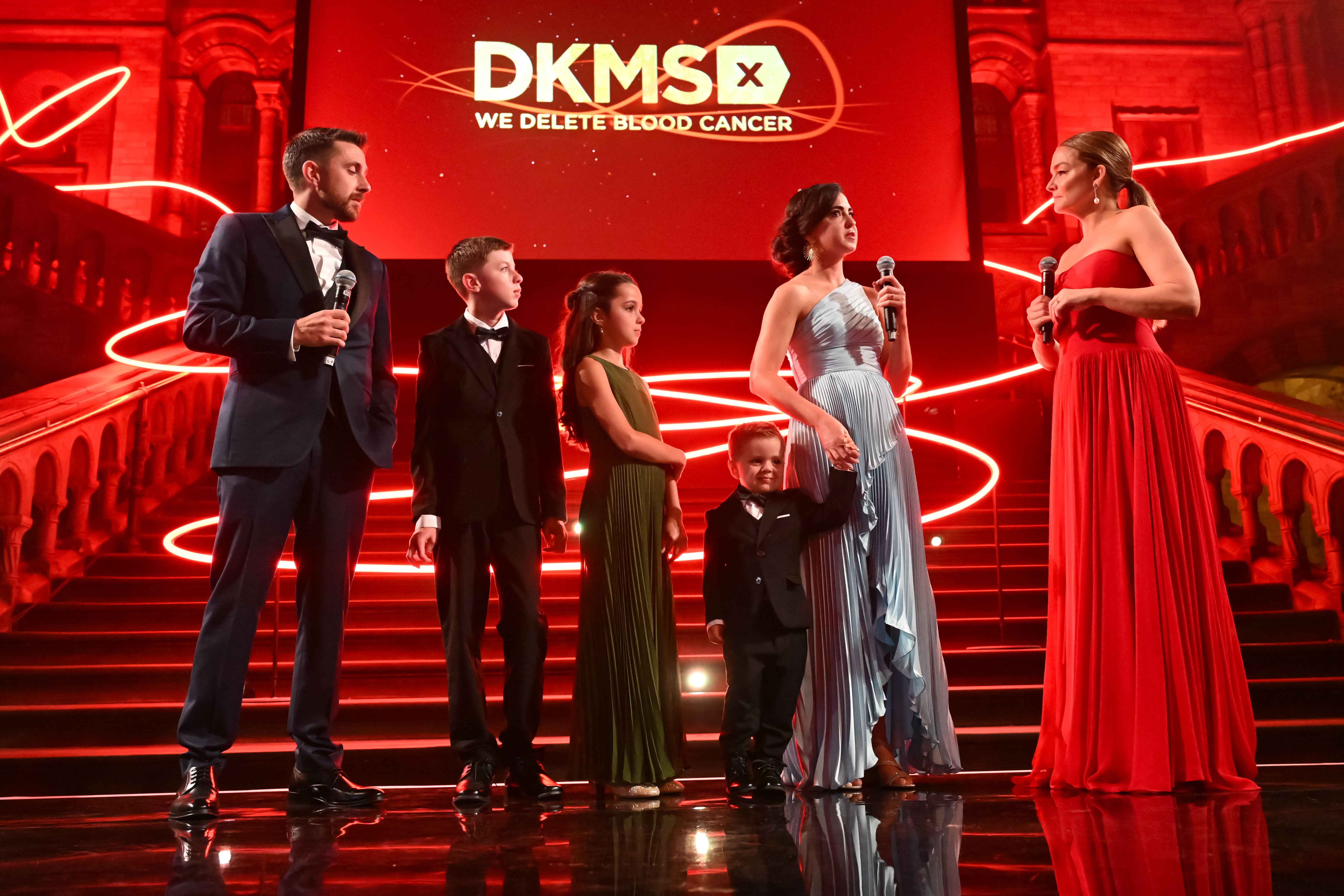 The Hoots family, including the patient Jonah Hoots, his parents and his two siblings, is standing on the left and speaking to Katharina Harf, who stands on the right side of the stage. All are dressed in elegant evening attire. Katharina Harf and the parents are holding microphones in their hands in front of the big staircase and the DKMS logo, speaking about their journey at the DKMS London Gals 2025.