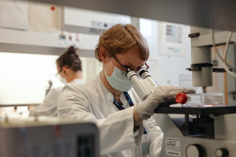 A female scientist wearing a lab coat, face mask, glasses, and gloves examines a sample through a microscope in a laboratory. She is focused on her work, holding the sample with her right hand. Another researcher is working in the background, highlighting a professional lab environment.