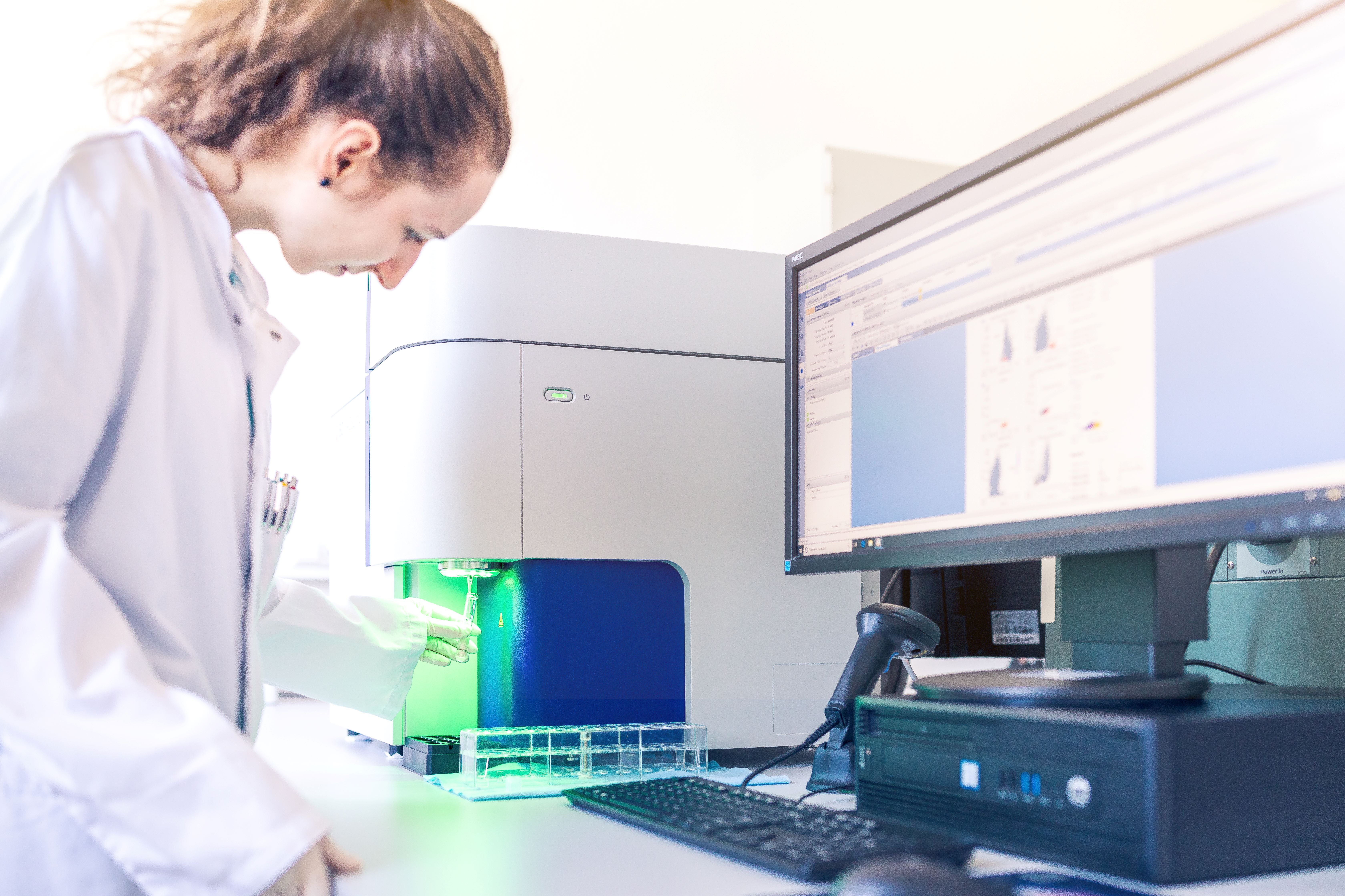 A young female scientist, wearing a white lab coat and gloves, is operating a flow cytometry machine in the DKMS Stem Cell Bank. A green light shines on the blood stem cell sample being analyzed, while a computer screen displays data and charts related to the quality of the blood stem cells.