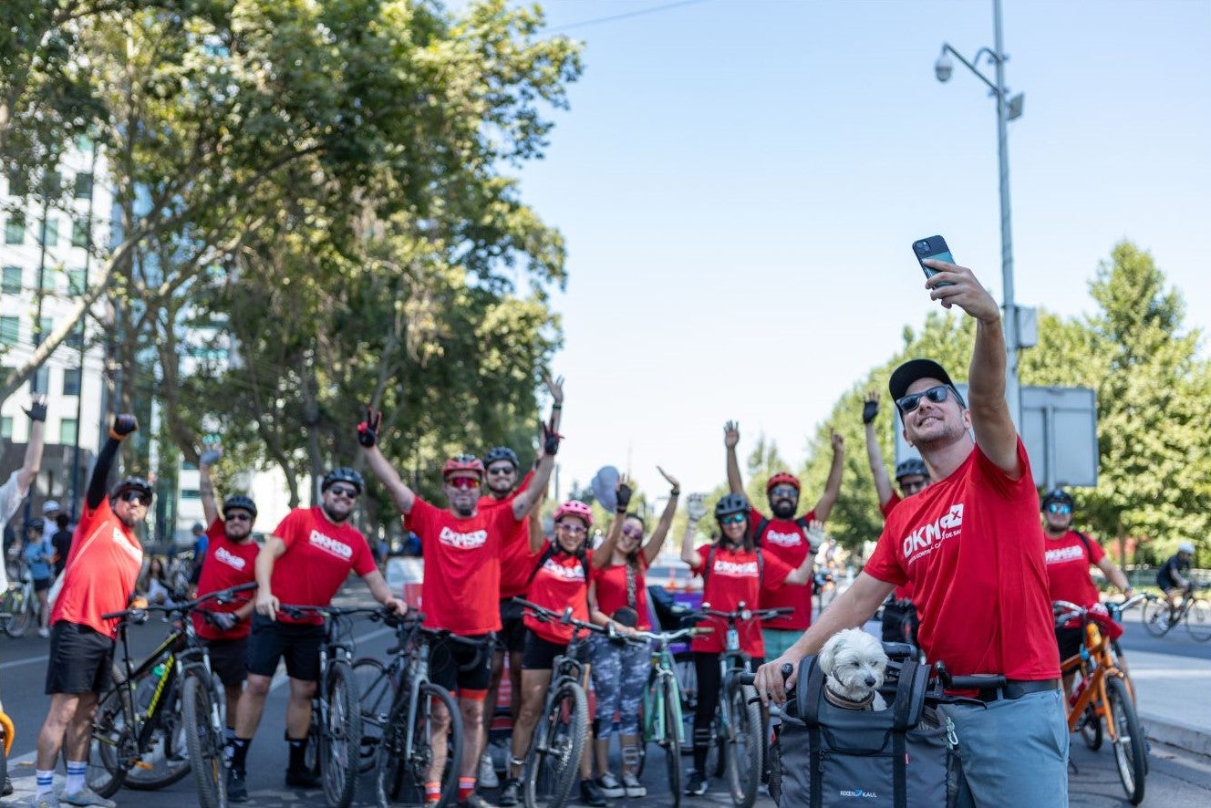 A group of cyclists, wearing red DKMS t-shirts, pose together outdoors. They are smiling, raising their arms, and standing with their bikes. In the front, the DKMS supporter Benedikt is taking a selfie with the group, while his small white dog Bruno sits in a basket on his bike.