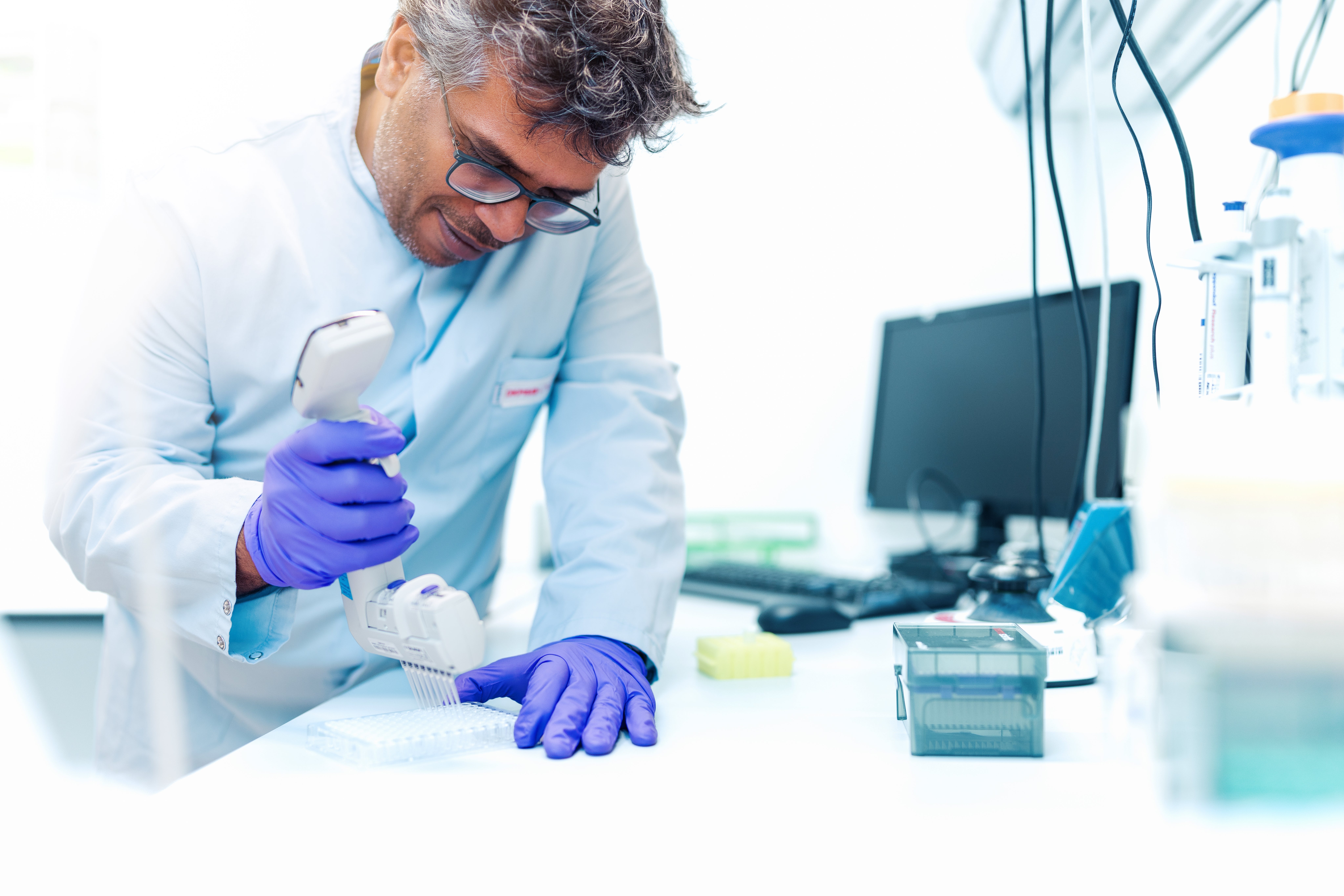 A male employee of the DKMS Life Science Lab, wearing a lab coat and purple gloves, uses a multichannel pipette to transfer samples into a well plate. Laboratory equipment and a computer are visible in the background.