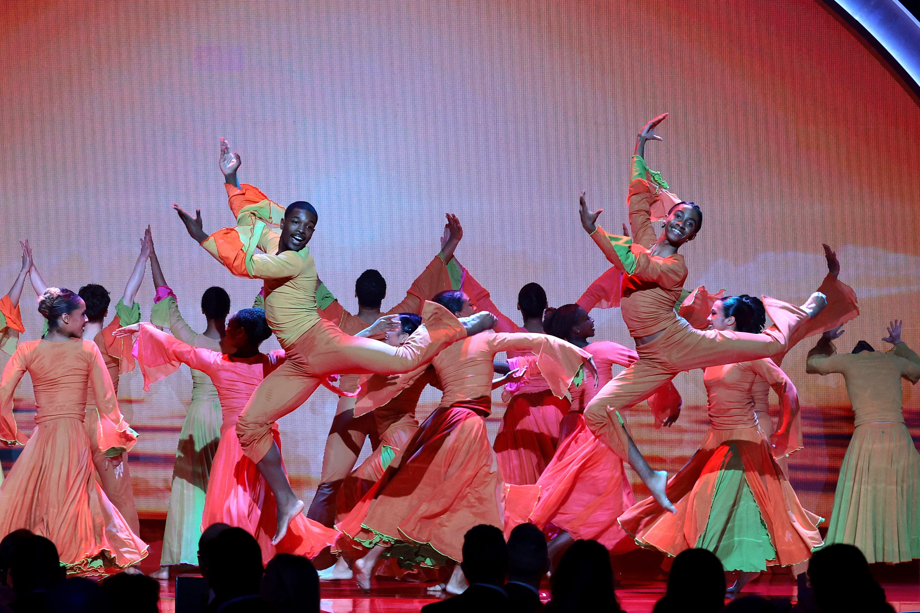 The Alvin Ailey Student Performance Group, wearing bright orange and pink costumes, perform an energetic dance performance on stage, with two dancers midair in dynamic jumps under colorful lighting.