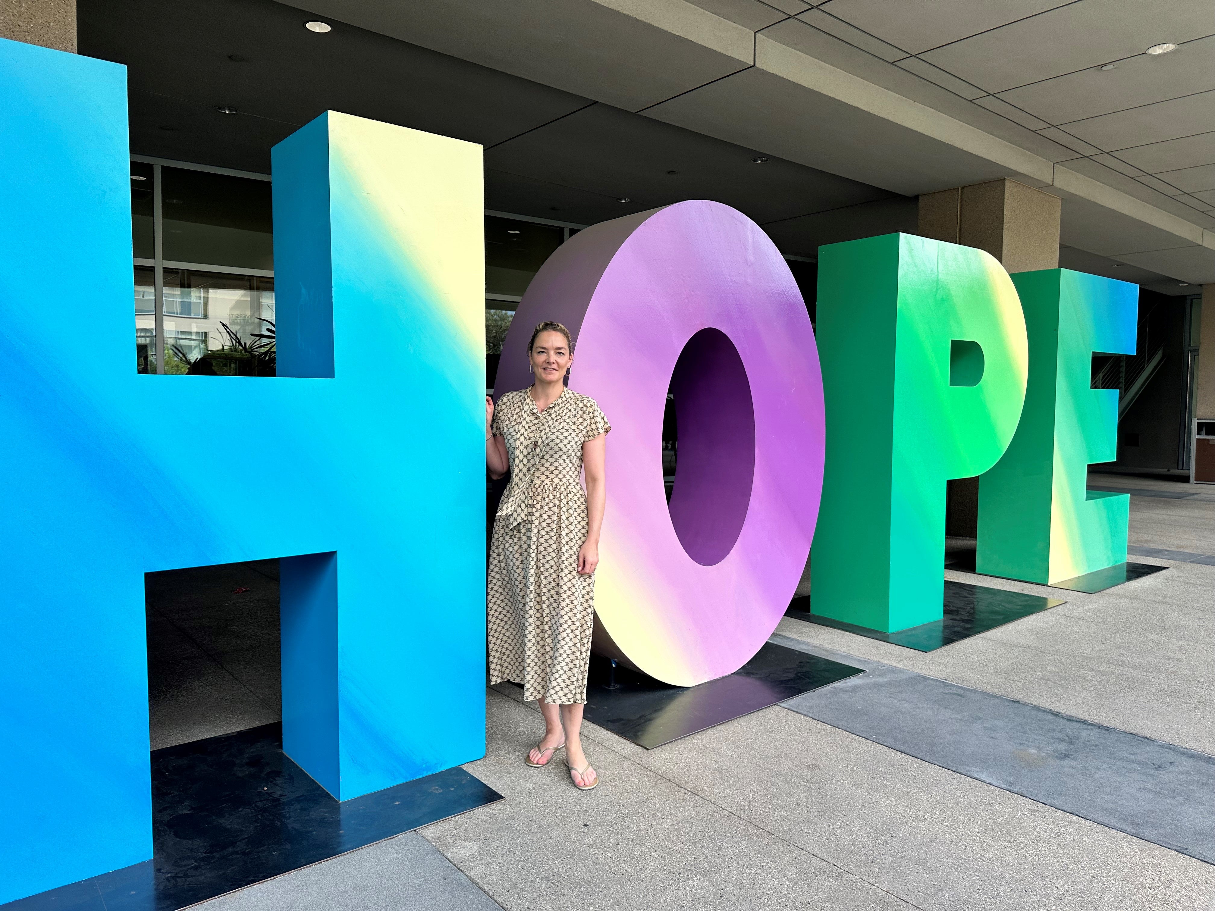 Katharina Harf stands next to a large, colorful sculpture spelling out the word 'HOPE' in gradient shades of blue, purple, green, and yellow. She is smiling and wearing a patterned light dress and sandals, standing in front of a modern building with a covered walkway.