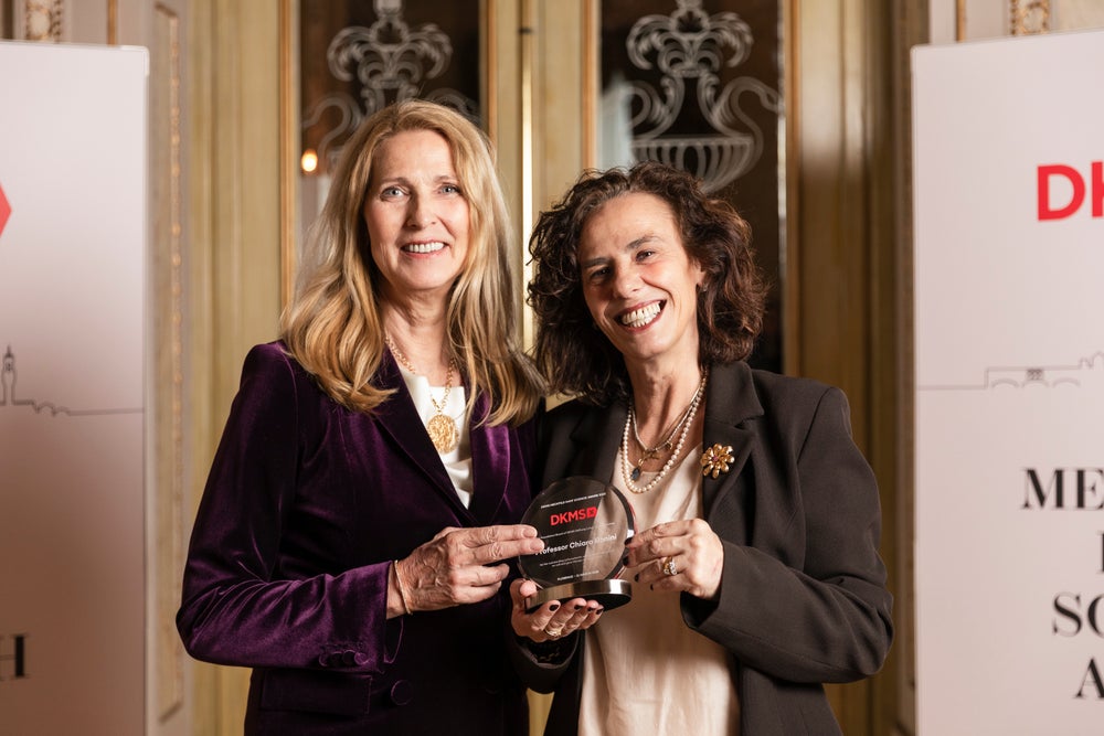 Dr. Elke Neujahr, CEO of DKMS, stands on the left next to Prof. Chiara Bonini, winner of the Mechtild Harf Science Award 2025, as they smile and hold the award together at the Mechtild Harf Science Award 2025 event. In the background DKMS banners and a large door are seen.