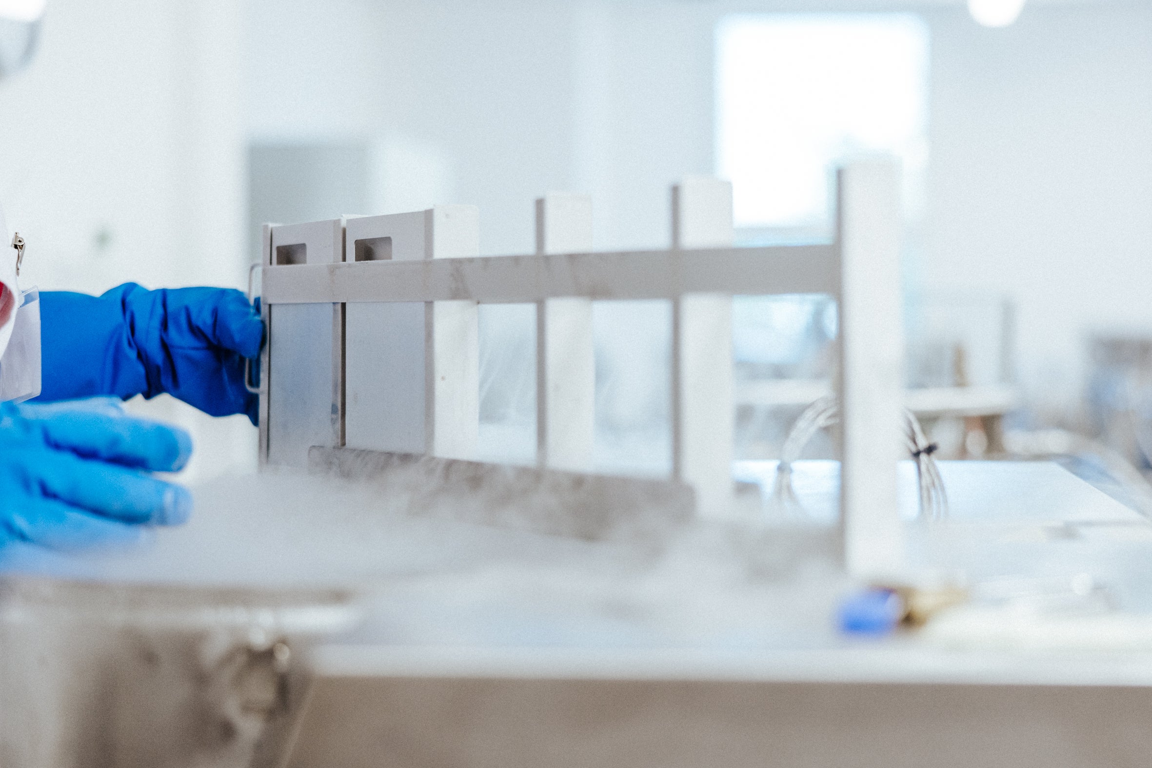 Close-up of hands of a laboratory employee with blue gloves in the DKMS Life Science Lab, who is inserting a cassette with ADCU into a predefined storage location on a rail. It's surrounded by vapor from liquid nitrogen.