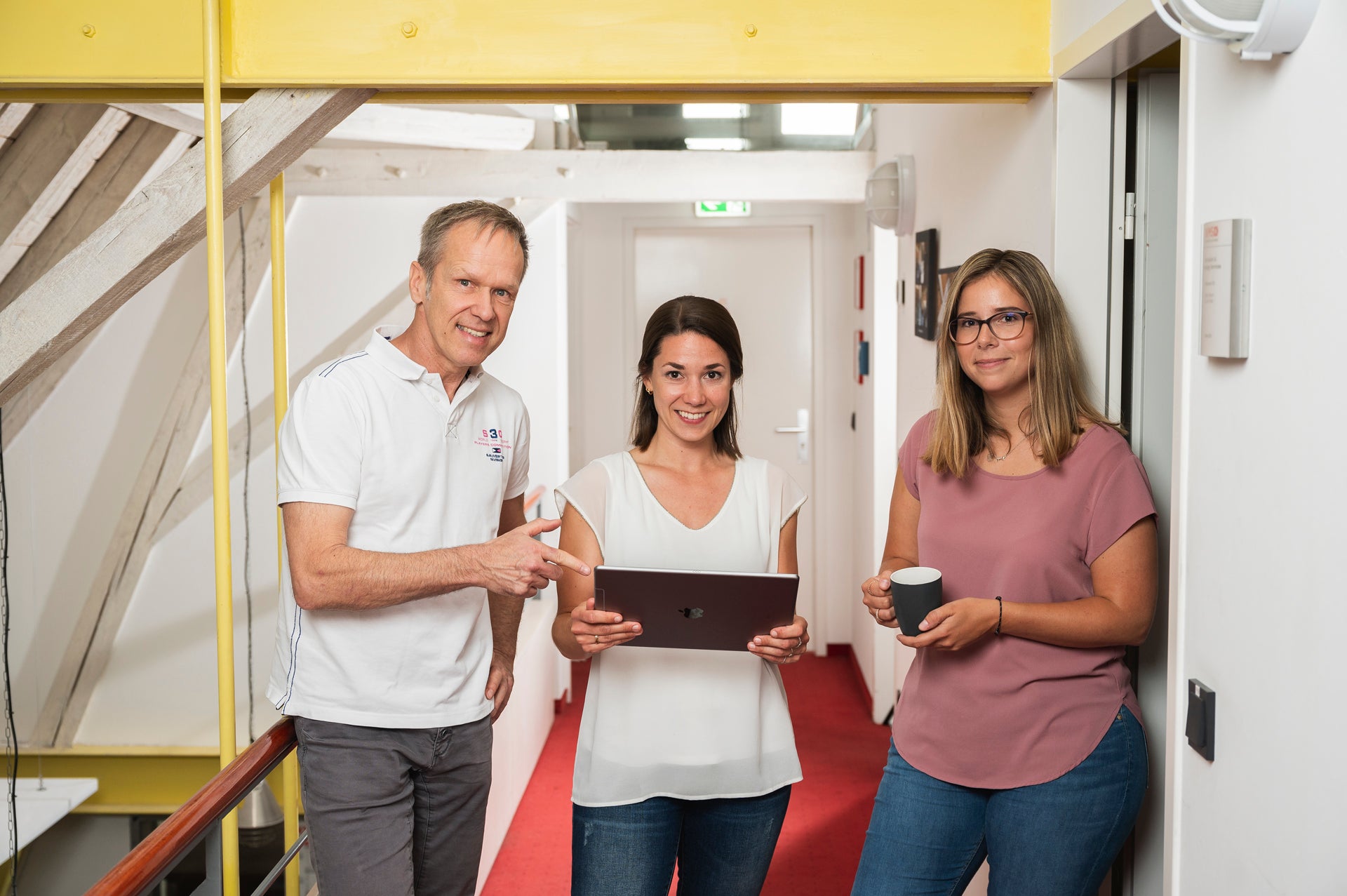 Three DKMS colleagues are standing together in a brigh hallway, smiling at the camera. The woman in the center is holding a laptop, while the man on the left points at the screen. The woman on the right is leaning against the wall, holding a coffee mug.