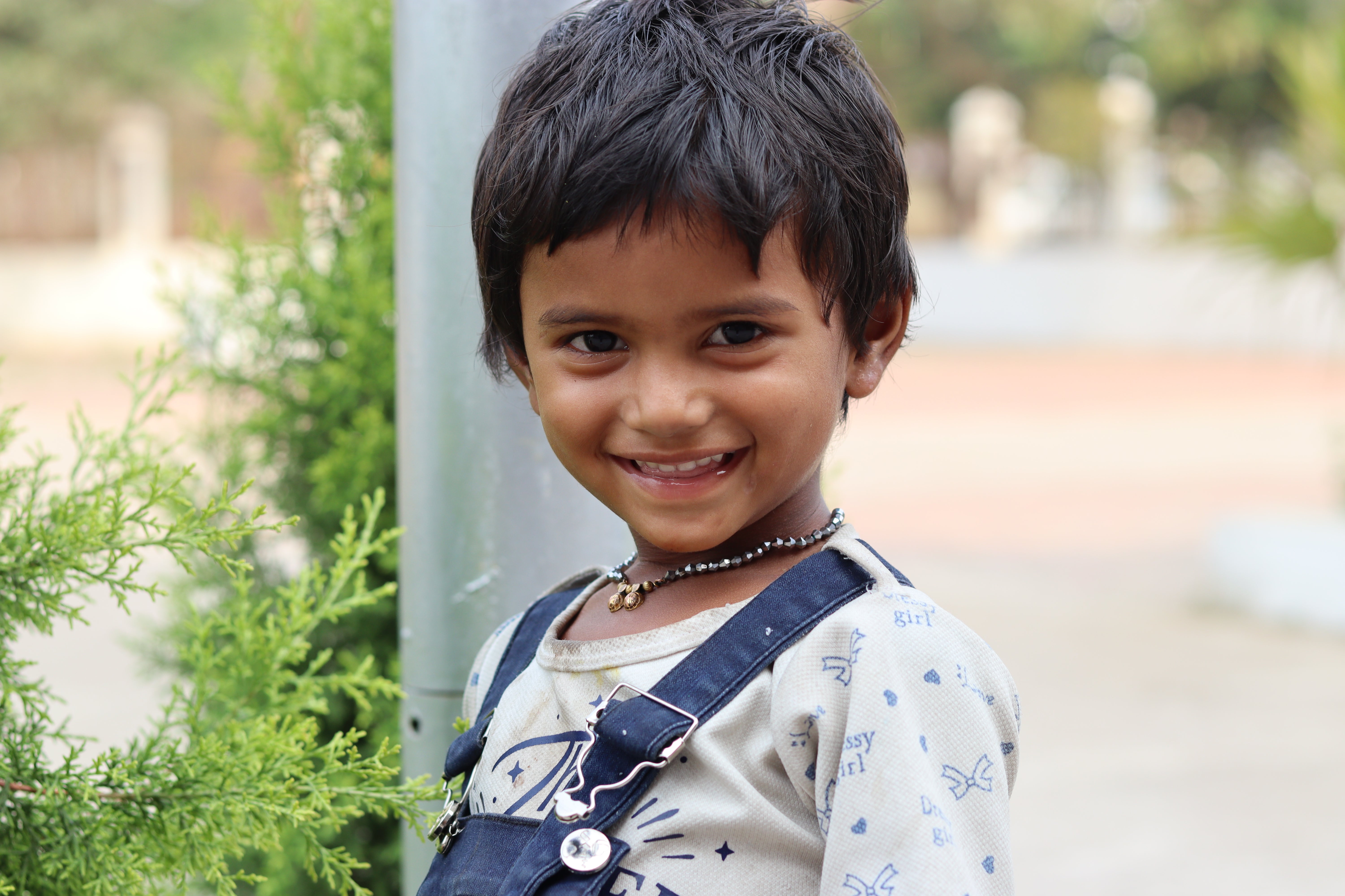 A young boy with black hair smiles warmly at the camera while standing outdoors next to green plants. The boy is wearing a light-colored shirt with printed patterns and denim overalls, and a beaded necklace. The background is softly blurred.