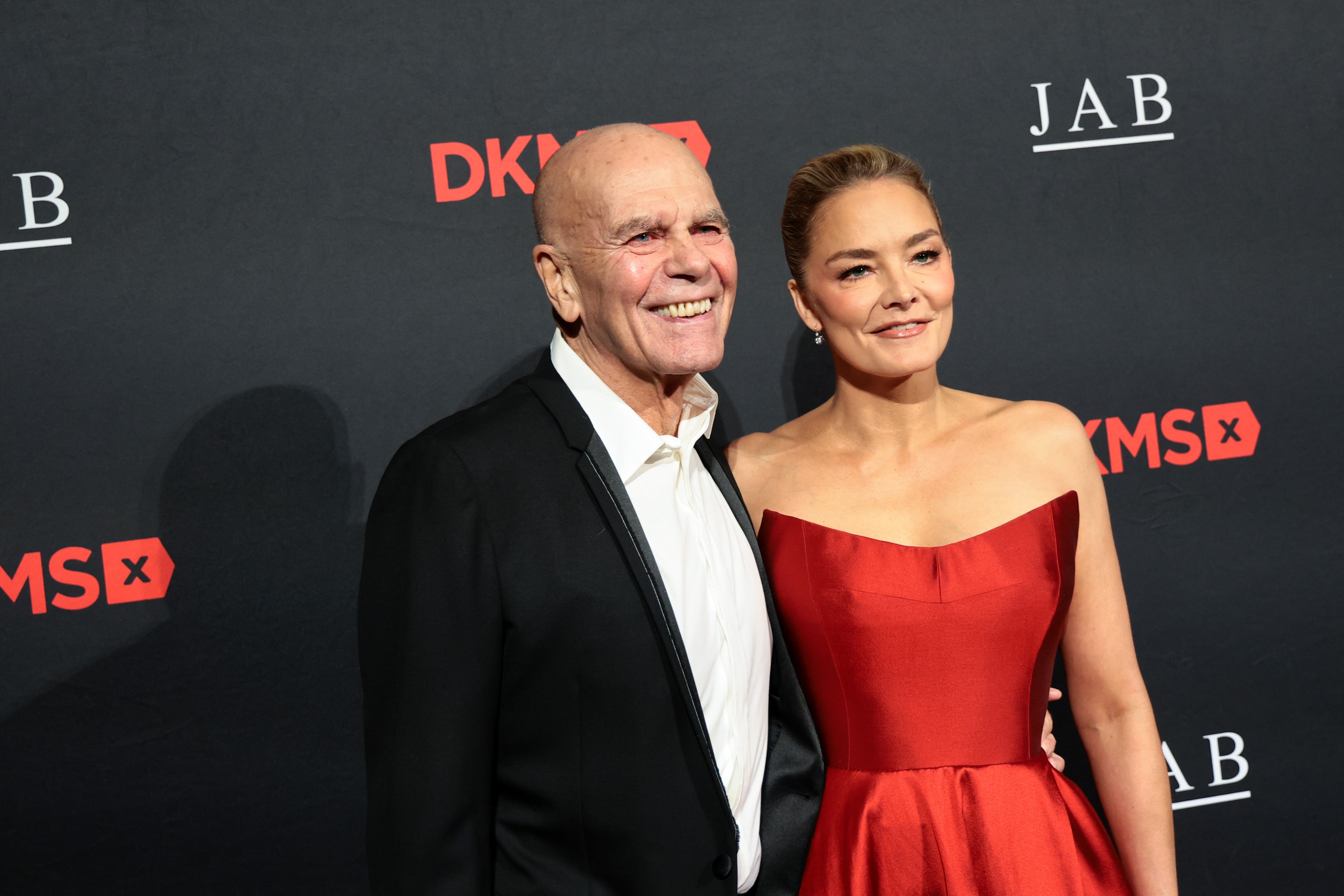 Peter Harf and Katharina Harf pose together on a red carpet in front of a black backdrop with DKMS and JAB logos. Peter Harf wears a black tuxedo, and Katharina Harf wears a strapless red gown. Both are smiling.