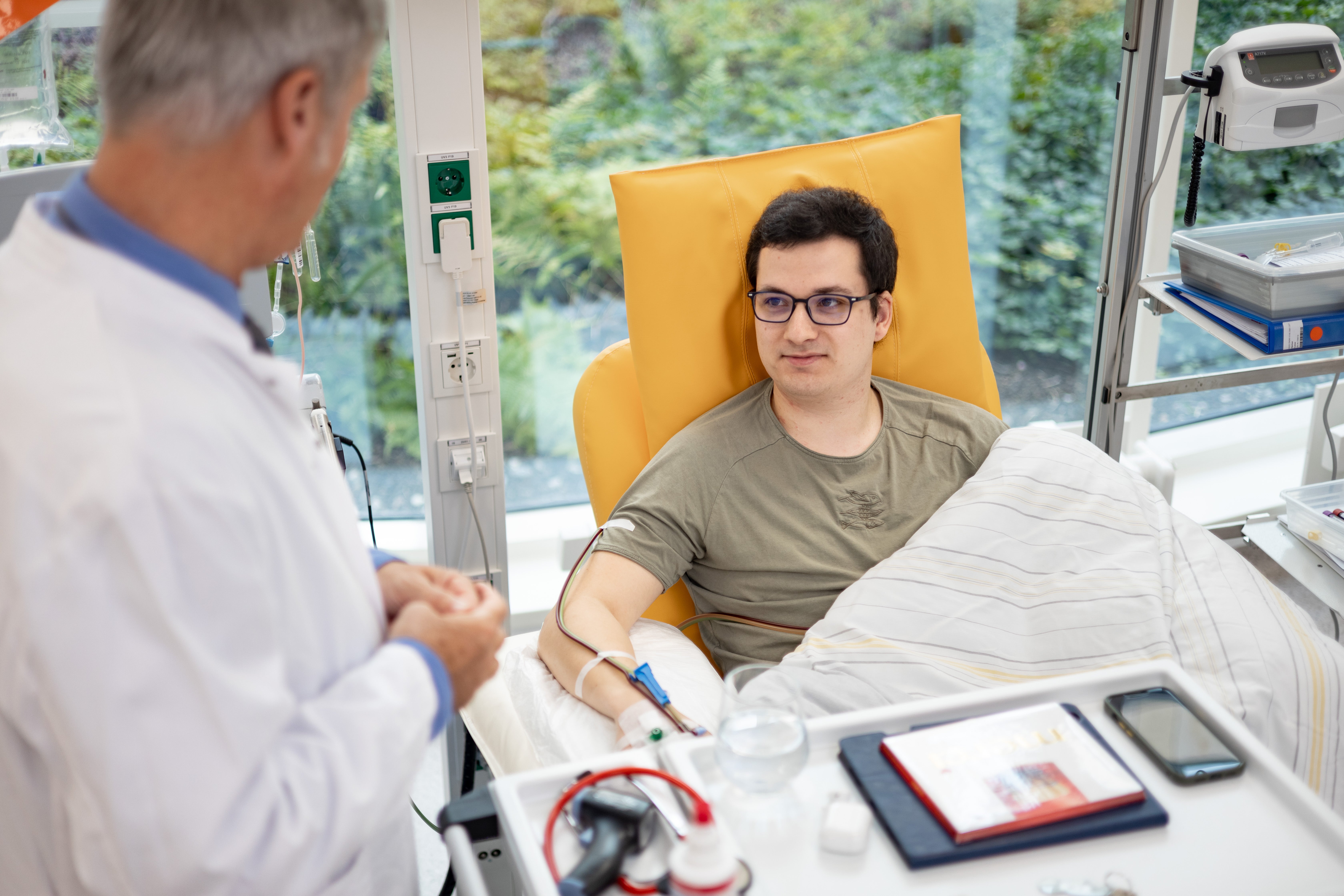 The young donor Lukas Hertzer is sitting in a yellow chair with a needle in his arm and connected to a apheresis tubing. He is the first donor whose cryopreserved stem cells were stored at the DKMS Stem Cell Bank. He is covered with a blanket and looking at Dr. Alexander Platz, the Medical Director of the Stem Cell Bank. In the background medical equipment and a big window with a view of the greenery outside are visible. 