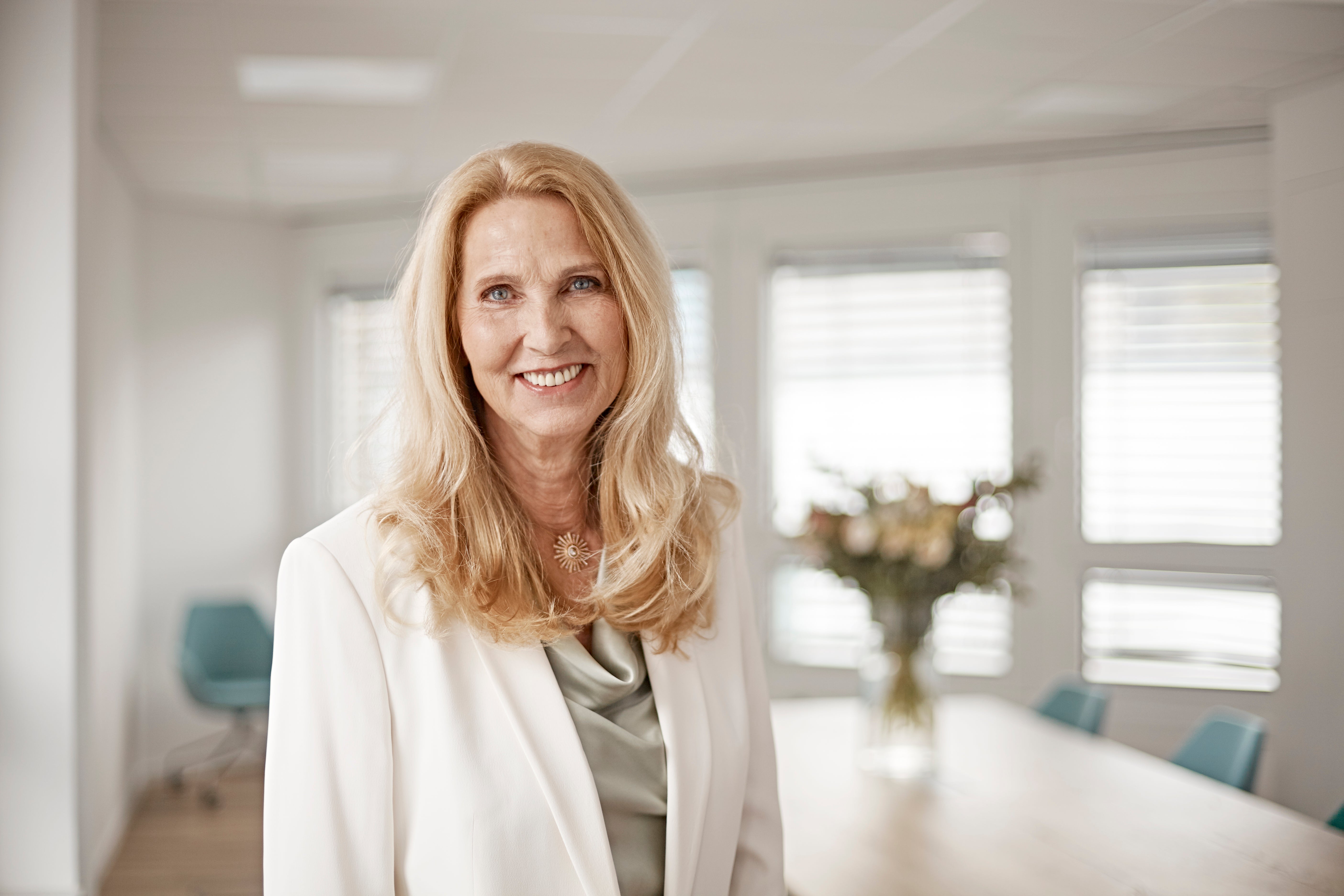 Portrait of Dr. Elke Neujahr, Global CEO of DKMS Group, standing in a bright office space. She is wearing a white blazer, a light sage satin blouse, a golden necklace and is smiling confidently at the camera. In the background is a bouquet of flowers on a office table, blue office chairs, and windows.