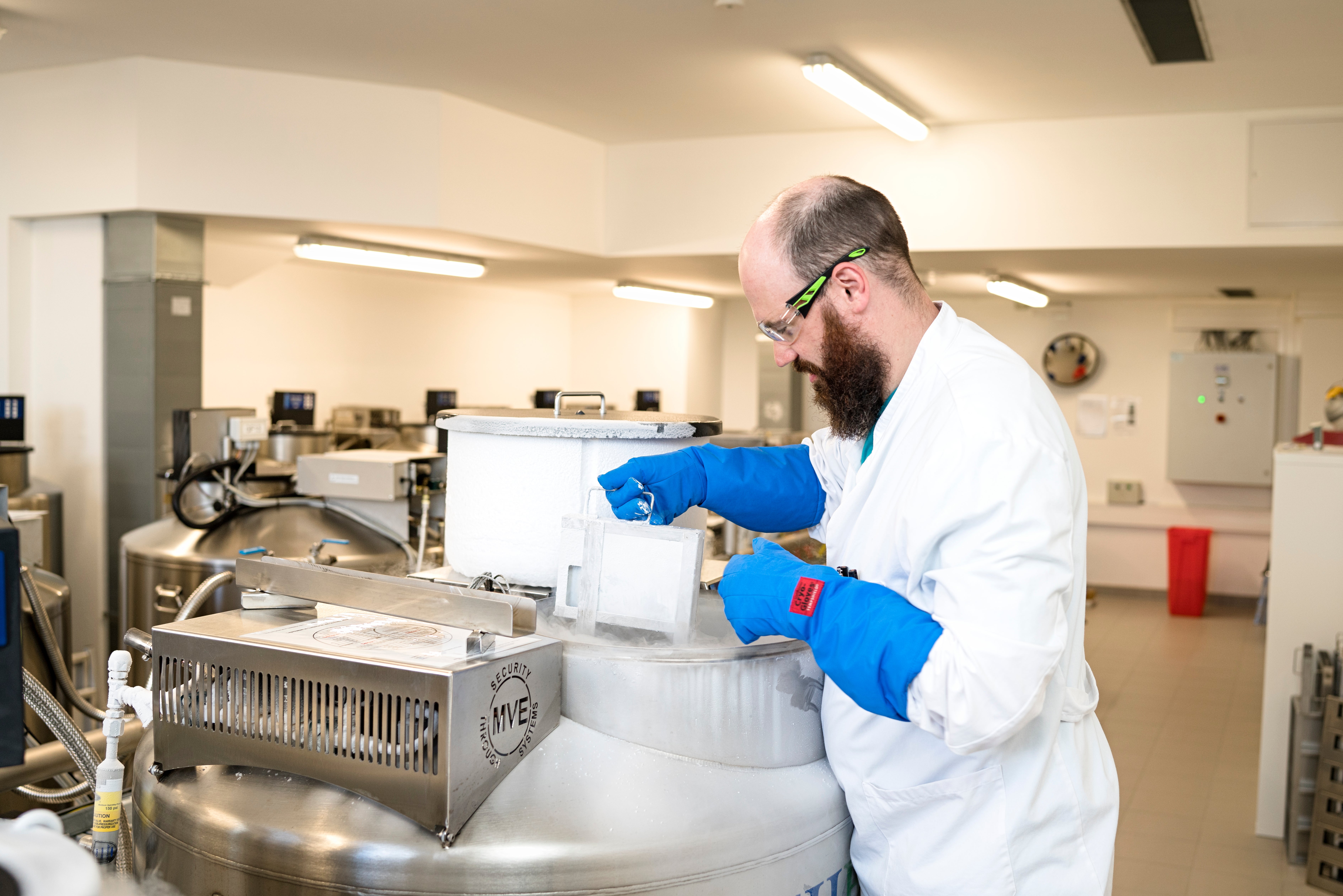 A male scientist at the DKMS Stem Cell Bank, wearing a white lab coat, safety goggles, and cryogenic gloves, is handling a storage rack with cryopreserved stem cell preparations, also called Adult Donor Cryopreserved Units (ADCUs) at a cryotank. More cryotanks can be seen in the background.