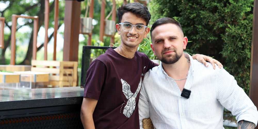 The thalassemia survivor Chirag and the stem cell donor Roman smiling at the camera and standing close together outdoors. Chirag on the left, is wearing glasses and a dark t-shirt, while Roman on the right wears a light striped shirt with a microphone clipped on. They have their arms around each other, posing for the photo in a green, outdoor setting.