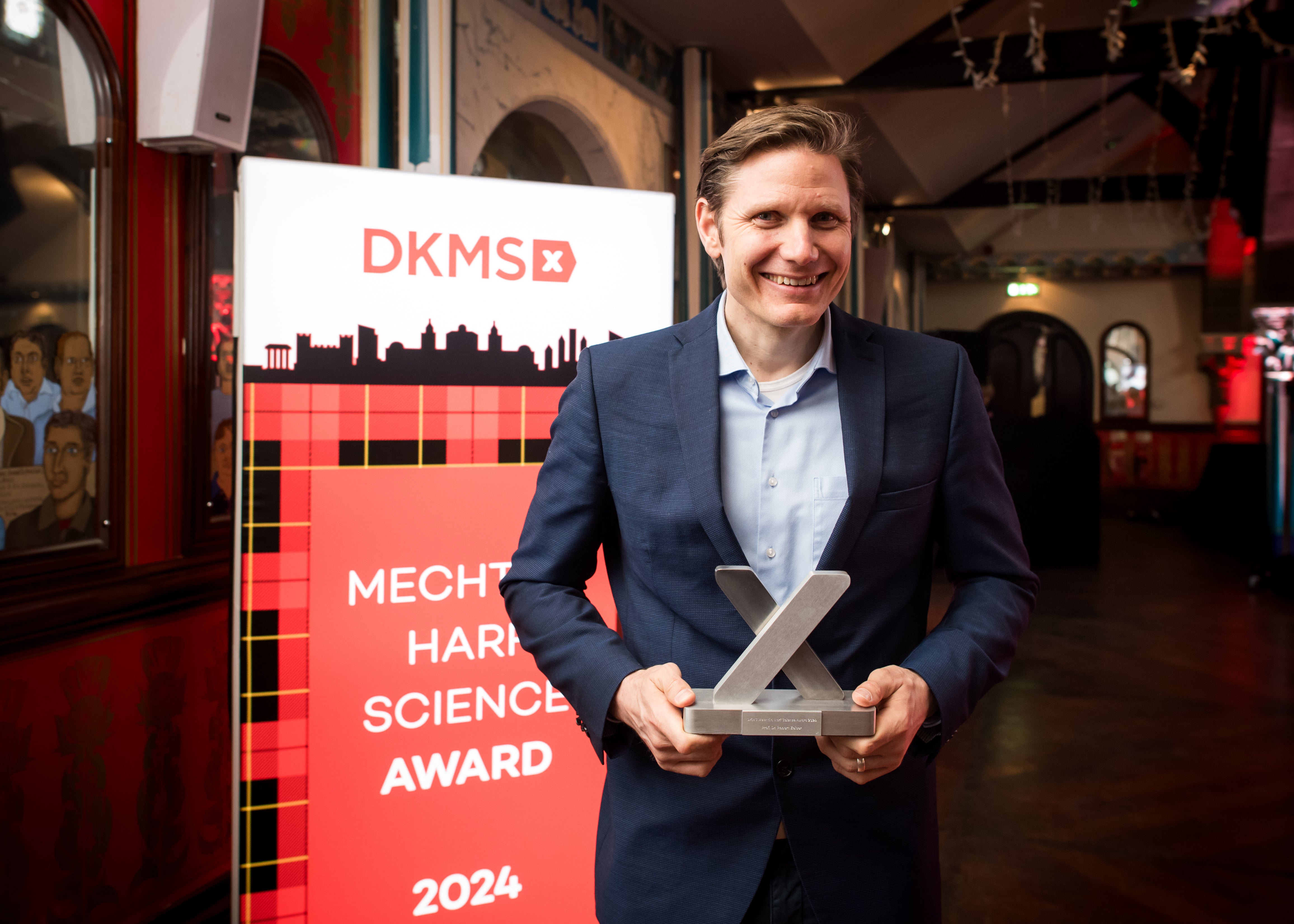 The winner of the Mechtild Harf Science Award Prof. Robert Zeiser, wearing a dark suit, holds the award trophy, in the shape of an x, and stands in front of a red DKMS banner that reads "Mechtild Harf Science Award 2024".