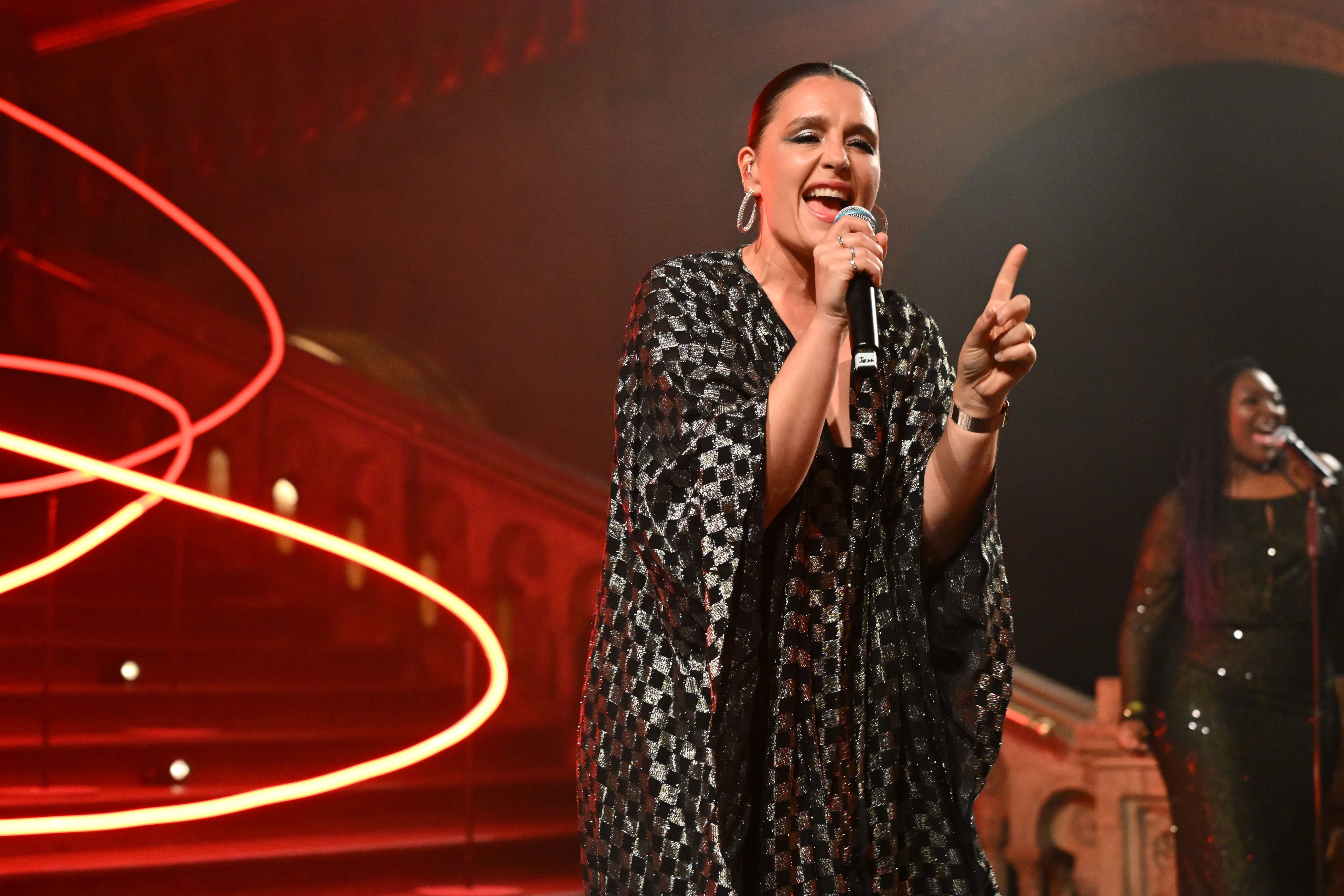 British singer and songwriter Jessie Ware performs on stage at the DKMS London Gala 2025, holding a microphone and wearing a sparkling black and silver dress with big silver creoles. In the background the big staircase, red lighting and a female background singer, who also sings in a microphone, is seen.