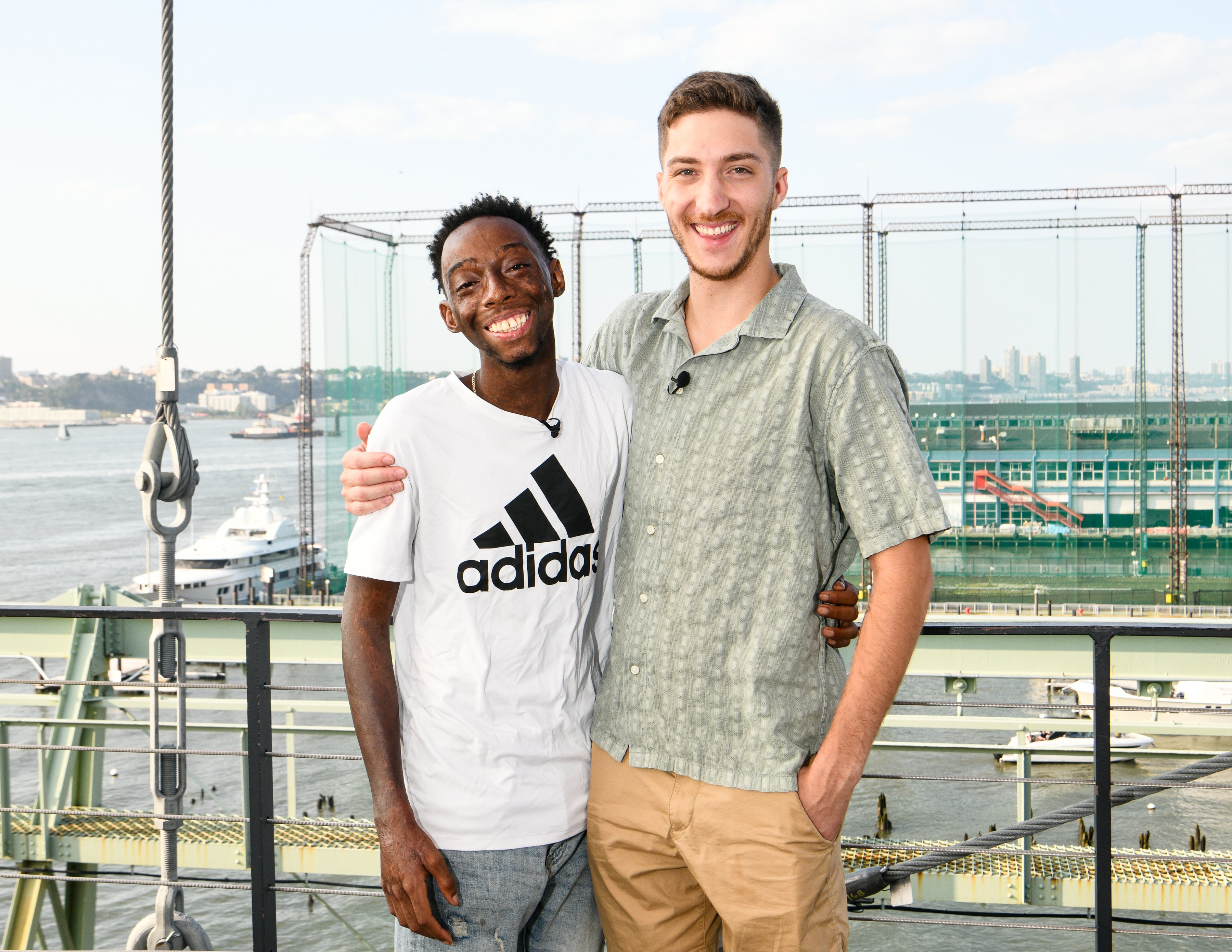 Two smiling young men, who are donor and patient, stand side by side on a platform with their arms around each other. The guy on the left wears a white Adidas t-shirt, the other one on the right a light green patterned shirt and beige shorts. In the background, a river, boats, and a city skyline are visible.