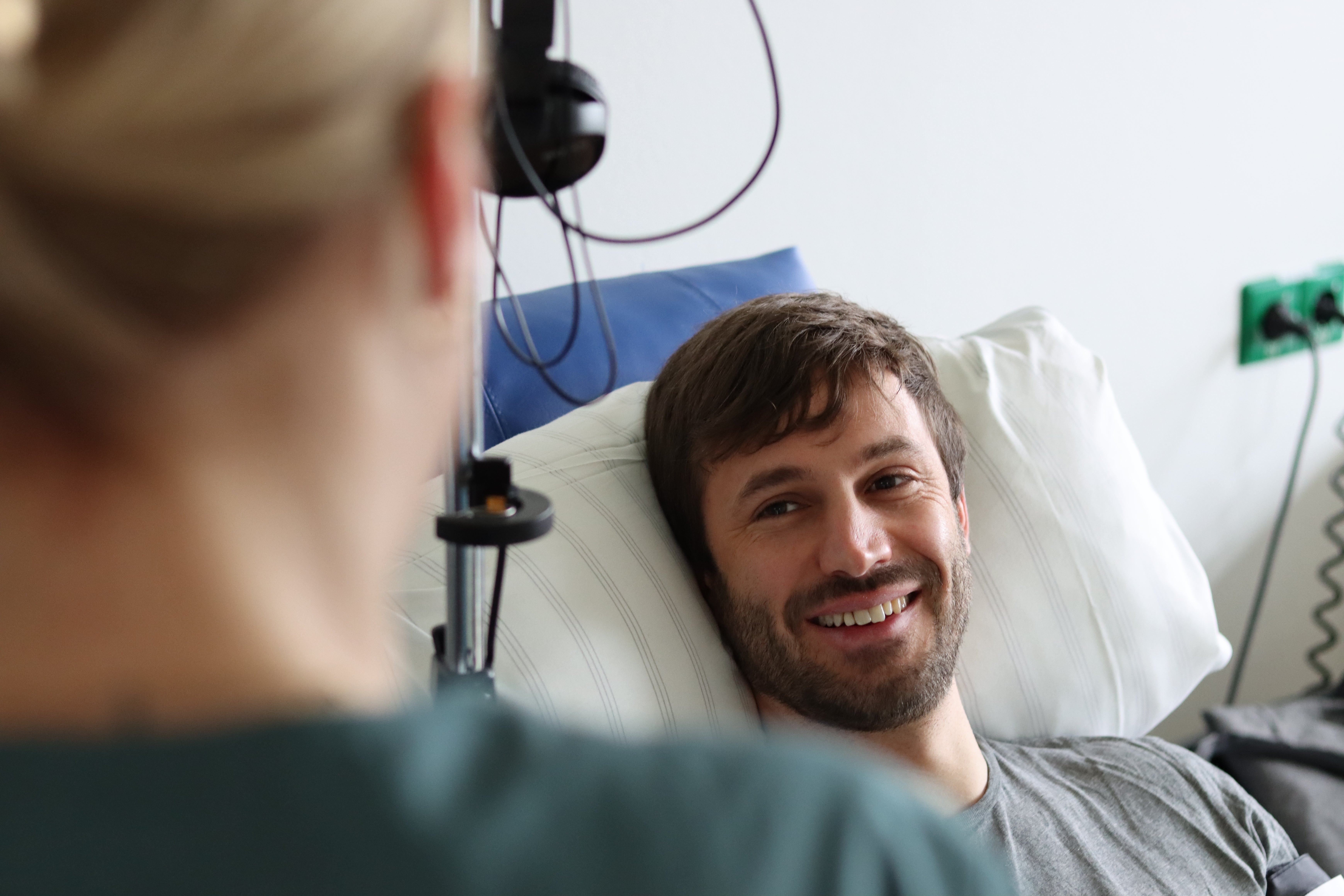 The donor Marvin is sitting in a chair for stem cell donation, smiling and looking at a woman in the foreground who is slightly out of focus.