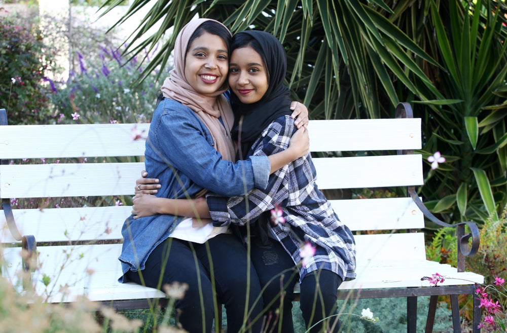 Two young women wearing hijabs are sitting on a white bench in a garden, smiling warmly and hugging each other. The woman on the left is a patient and wears a denim jacket and a beige hijab, the other woman on the right wears a plaid shirt and a black hijab. Both wear black trousers. Lush green plants and colorful flowers surround them.