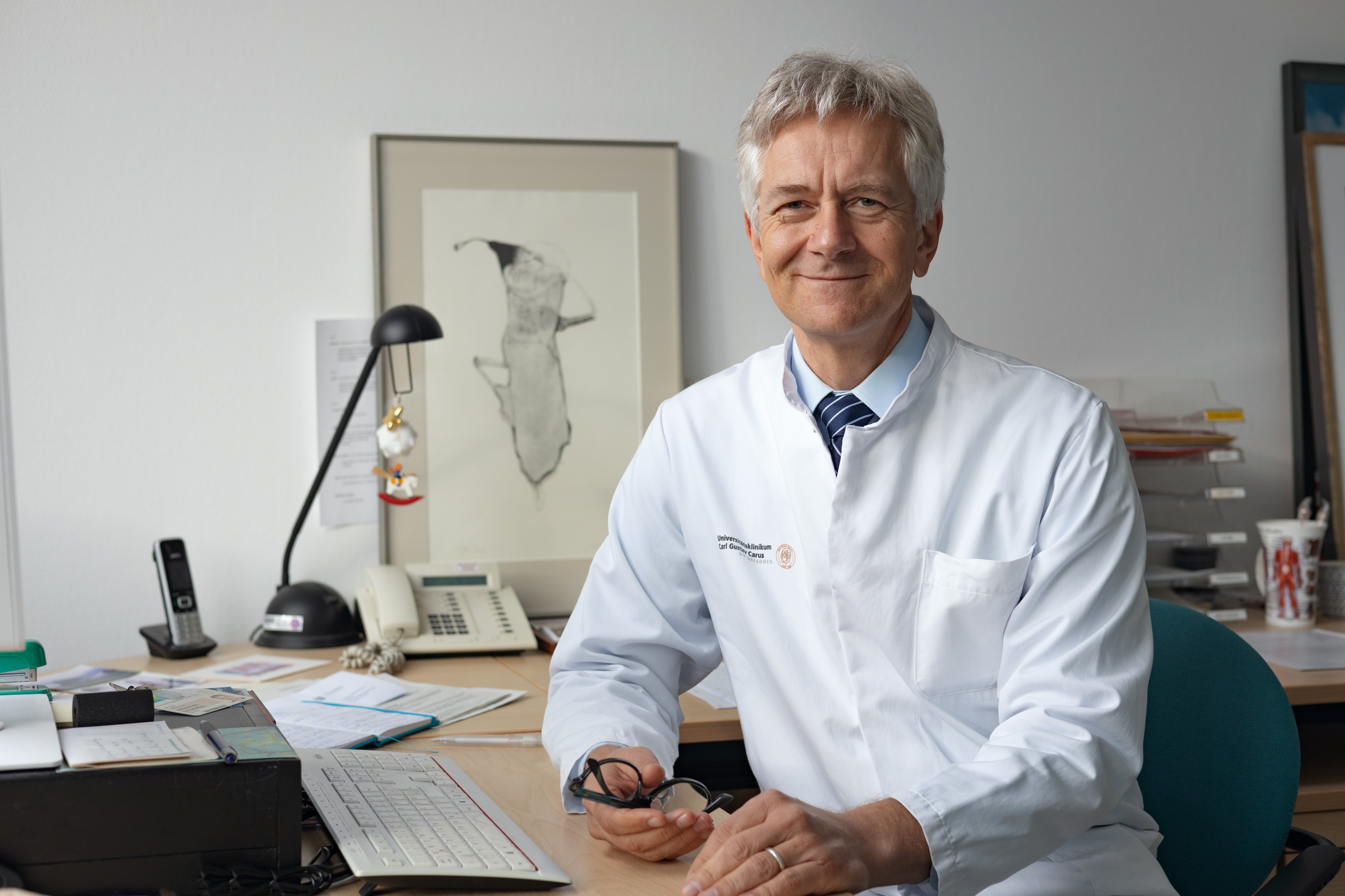 Prof. Dr. Johannes Schetelig wearing a white coat smiles gently at the camera and sitting at a desk in an office. He is holding glasses in his right hand, with papers, a keyboard, and a telephone visible on the desk behind him.