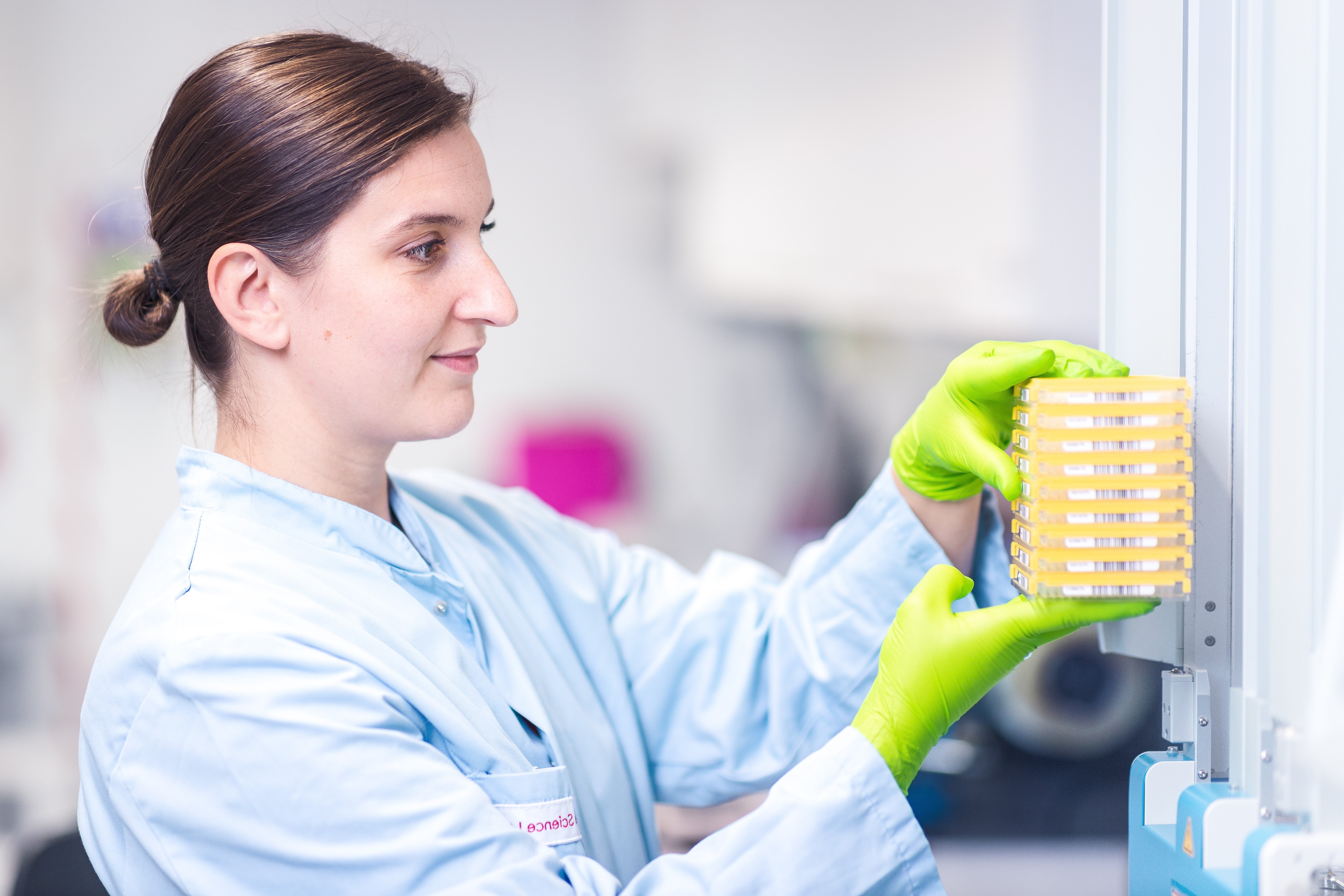 A female employee at the DKMS Life Science Lab, with brown hair in a bun, wearing a light blue lab coat and green protective gloves is working in the laboratory. She is holding a yellow stack of sample containers and placing them into a machine. The background is blurred.