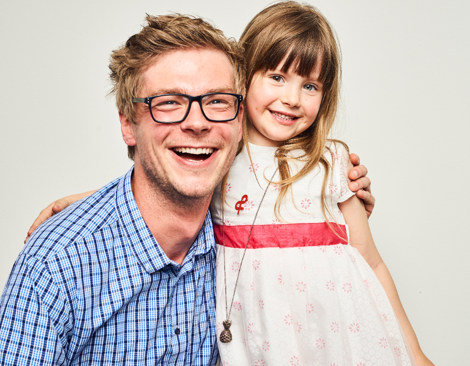 A smiling man wearing black glasses and a checkered blue shirt is sitting to the left of a little girl in a white dress with a red sash. Both are happily looking at the camera with their arms around each other.