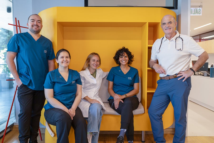 Five Chilean medical professionals pose together in front of a bright yellow wall structure inside a modern clinic in the Donor Center in Chile. The three women are sitting in the middel, and the two men are standing on either side. They are wearing white and dark blue medical scrubs and shirts, smiling warmly.