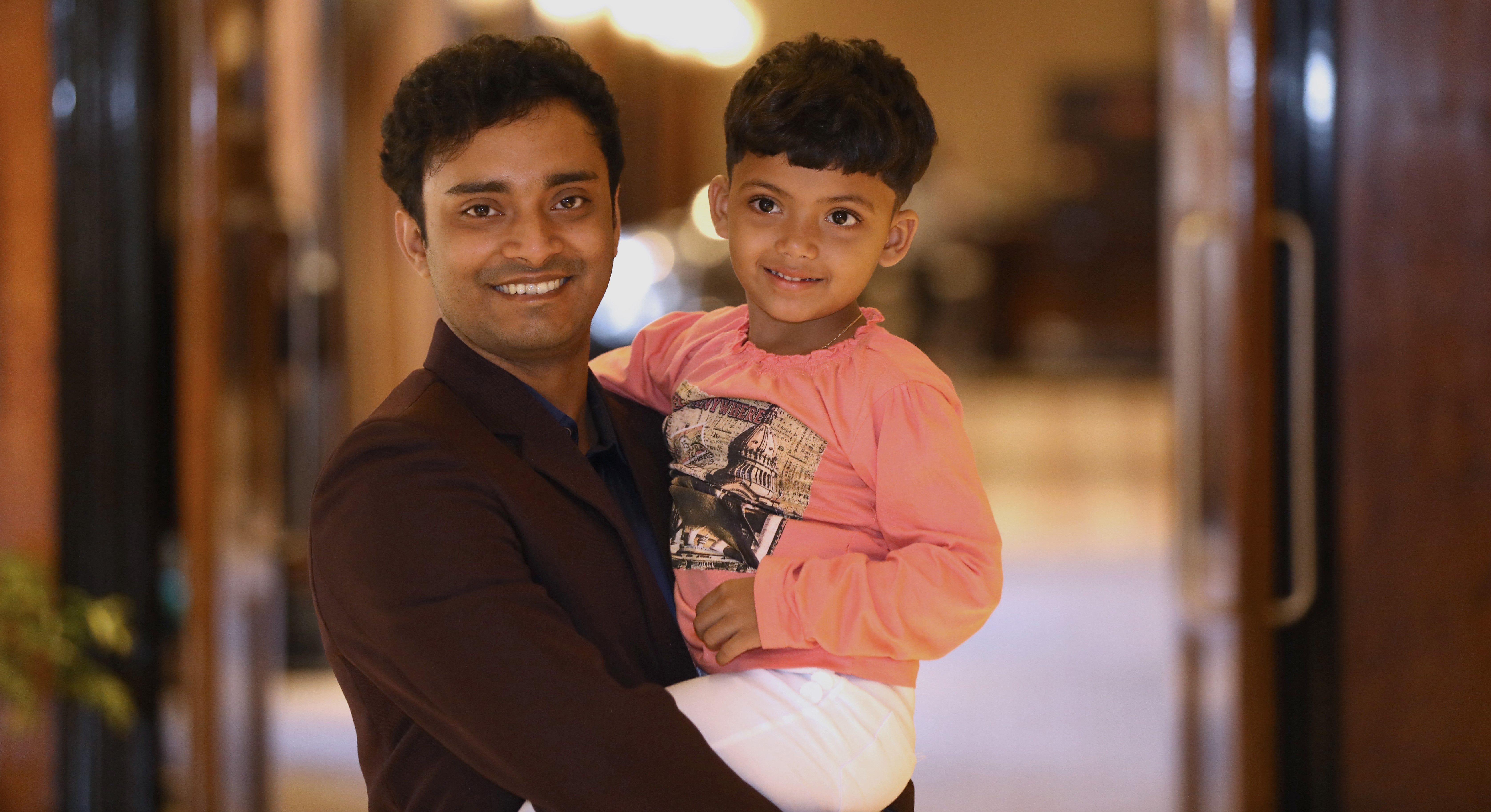 A smiling man holds a young boy, who are donor and patient, in his arms. The man is wearing a dark shirt, and the boy is dressed in a pink long-sleeve shirt and white pants. Both are looking at the camera with warm, happy expressions. The softly lit background suggests a cozy indoor setting.