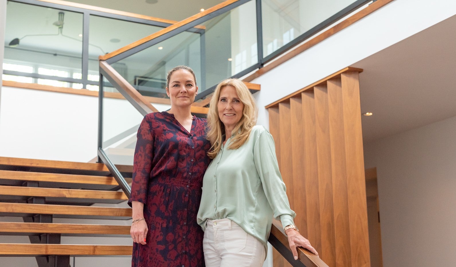 Katharina Harf and Elke Neujahr standing side by side on a modern staircase in a bright interior. The steps are made of wood and the banister is made of glass. Katharina Harf wears a dark red and navy dress, and Elke Neujahr a light green blouse with white trousers. They are smiling confidently at the camera.