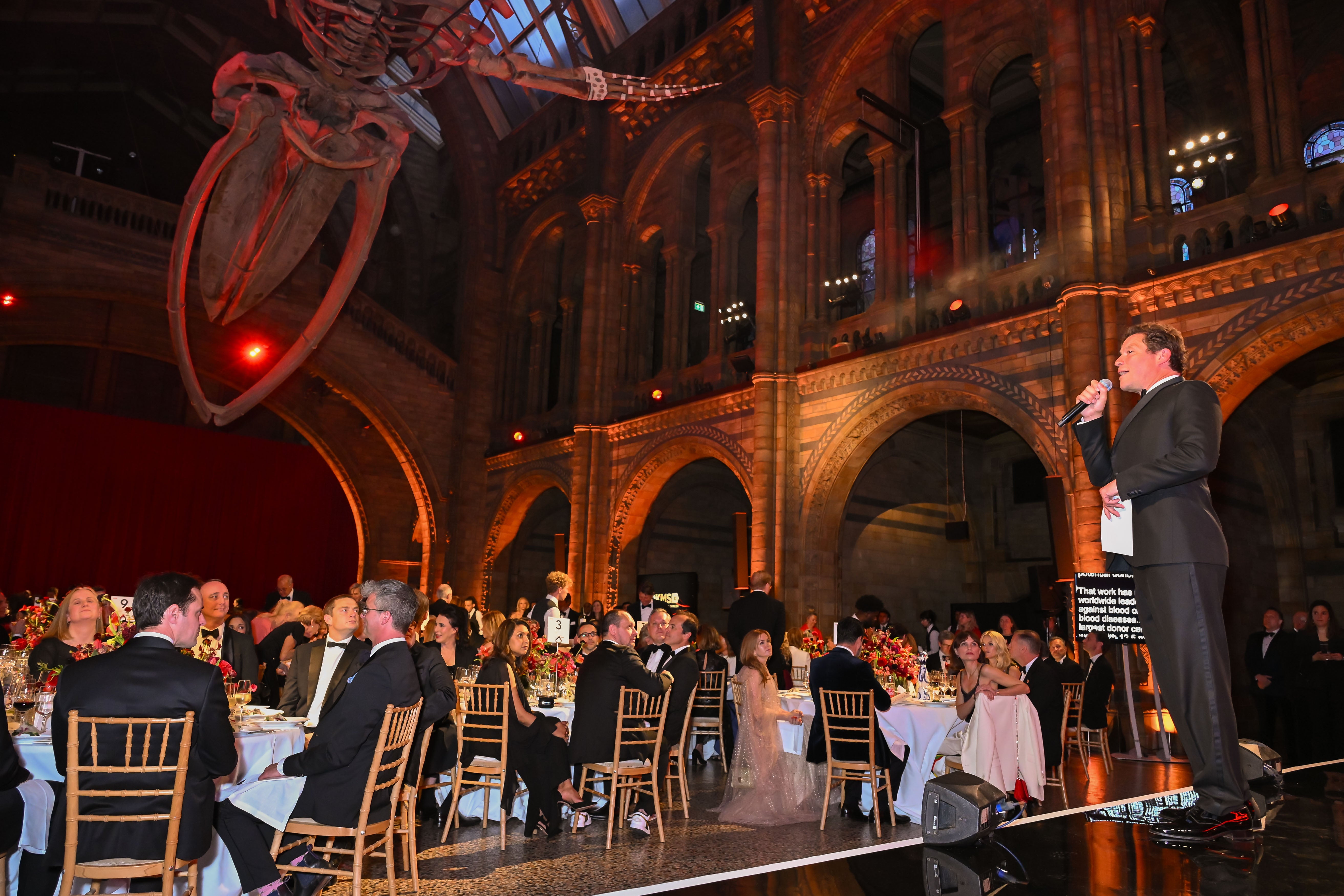 The gala host and actor Dominic West stands on stage holding a microphone, addressing a seated audience at the DKMS London Gala 2025 inside The Natural History Museum with high arches and a large whale skeleton suspended above.
