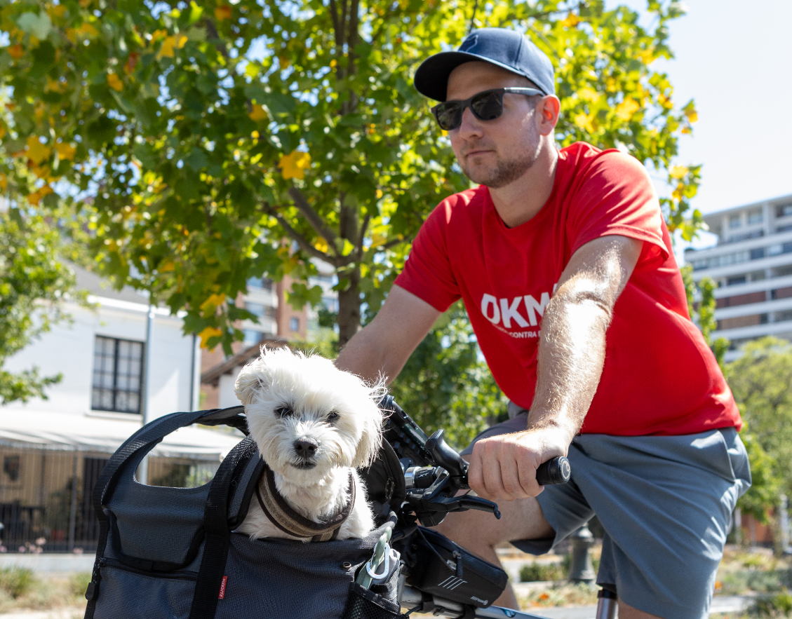 The DKMS supporter Benedikt, wearing sunglasses, a cap, and a red DKMS t-shirt, rides a bicycle outdoors. His small white dog Bruno sits in a front bike basket, looking at the camera. Trees and buildings are visible in the background.