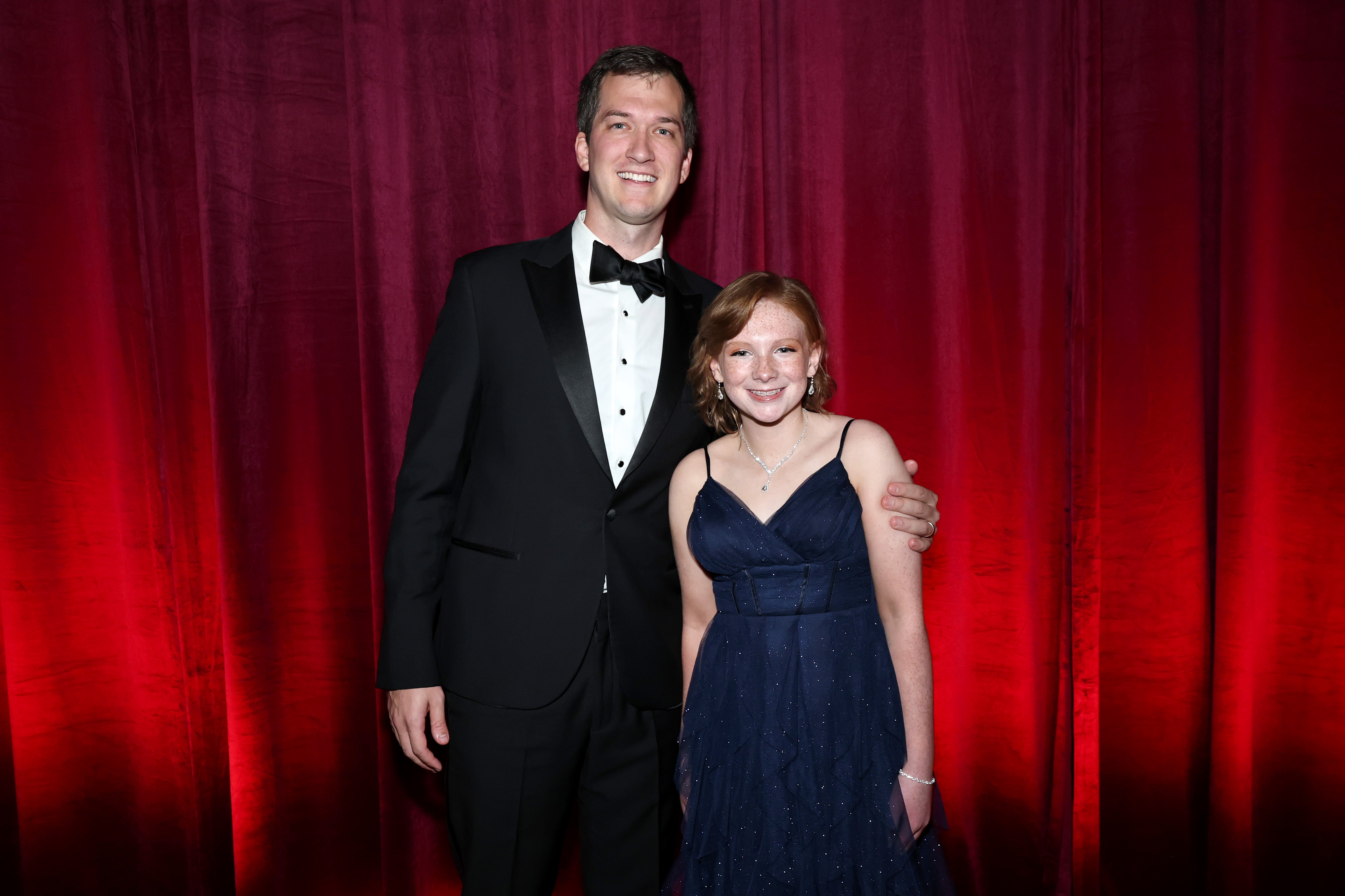 The DKMS donor William Dossett, wearing a black tuxedo, stands beside the 13-year-old acute myeloid leukemia survivor Madison Adams, wearing a navy blue gown. They are both smiling in front of a red curtain backdrop at the DKMS New York Gala 2025.