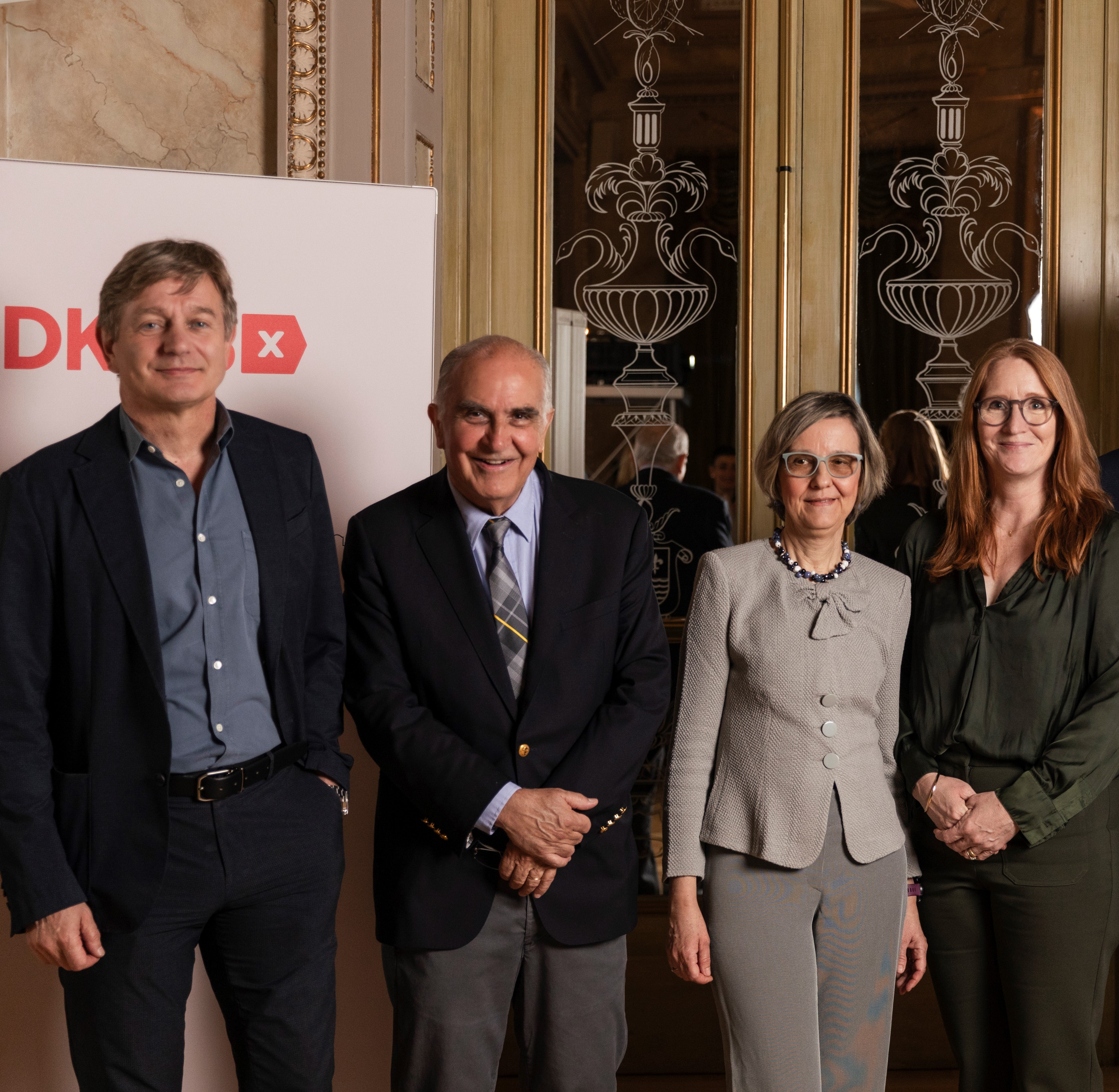 Four members of the DKMS Medical Council stand together in front of a decorative mirror background at the Mechtild Harf Science Award 2025. From the left you can see Prof. Dr. Marcel van den Brink, Dr. Marcelo Fernández-Viña, Prof. Dr. Katharina Fleischhauer, and Prof. Dr. Emma Morris. They are dressed in business attire and smiling at the camera, with DKMS signage partially visible on the left side.