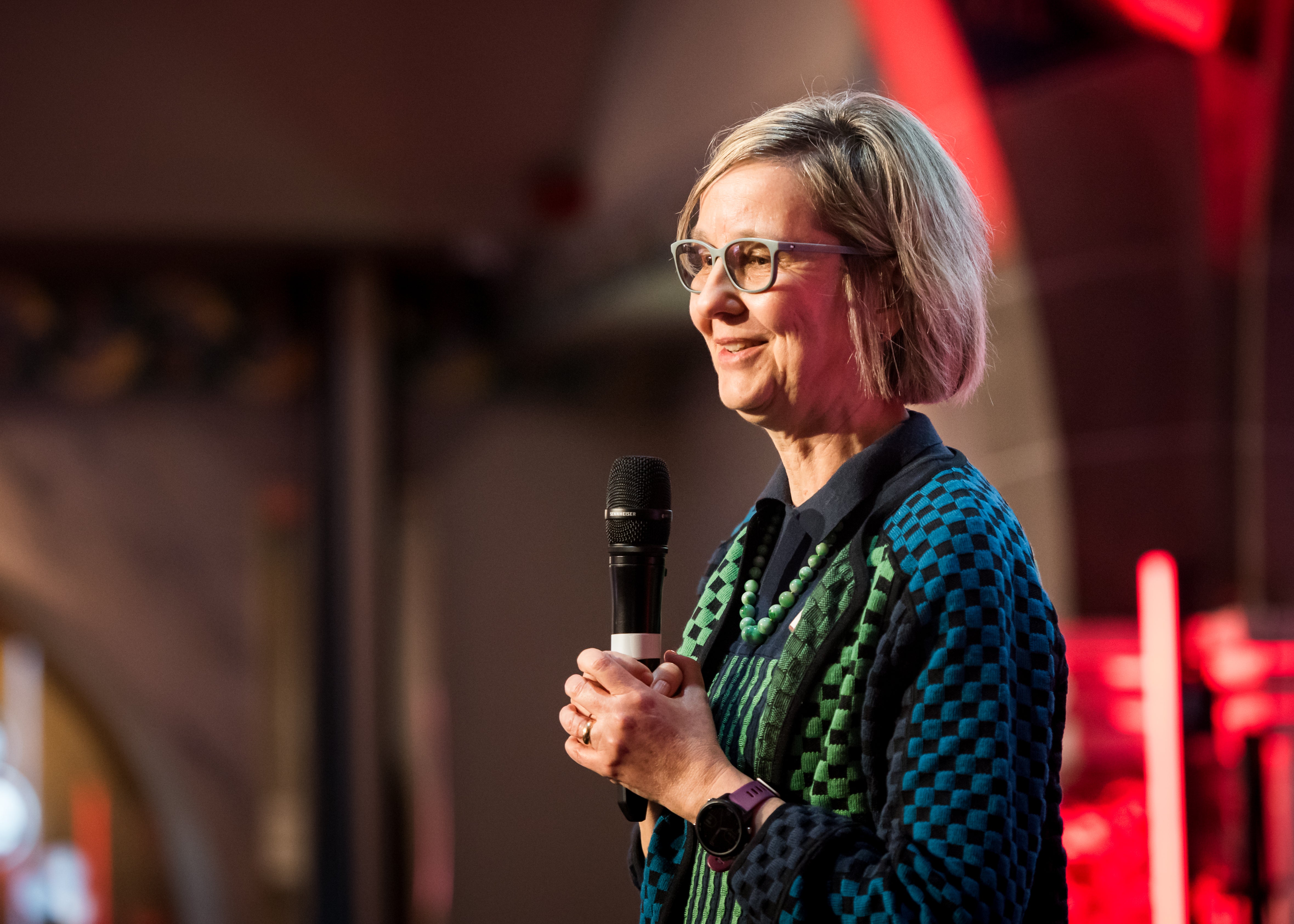 Prof. Katharina Fleichhauer, wearing glasses, a dark patterned jacket, and a necklace, holds a microphone in her hands while speaking on stage at the Mechtild Harf Science Award 2024. The background is softly lit with red lighting.