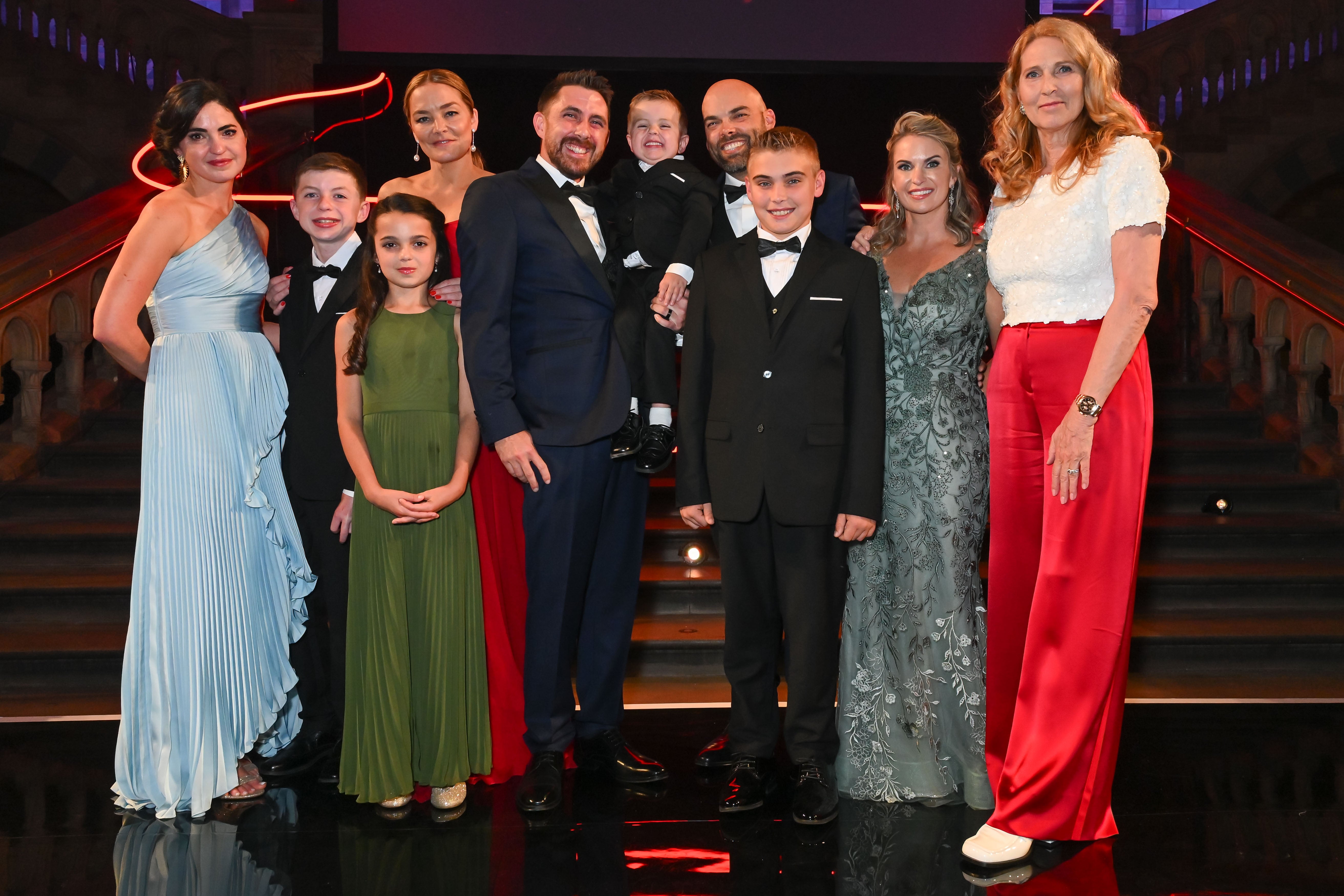 A group photo of ten people at the DKMS London Gala 2025 showing the patient Nate Hoots with his family, his donor Nate, Dr. Elke Neujahr and Katharina Harf. They are all dressed in elegant evening attire, smiling in front of a grand staircase.
