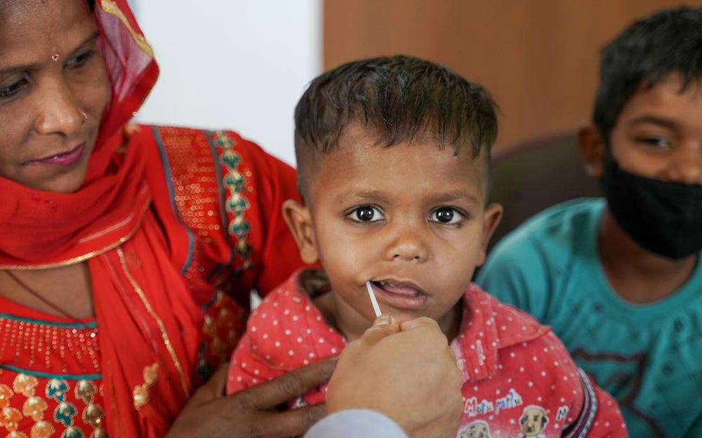 A young boy in a red shirt, who is a thalassemia patient at thal camp in India,  is being swabbed on the inside of the cheek, likely for a stem cell registration. The boy is looking in the camera and is sitting on a woman's lap, who is dressed in a vibrant red traditional outfit. Another boy wearing a turquoise shirt and black face mask is visible in the background.
