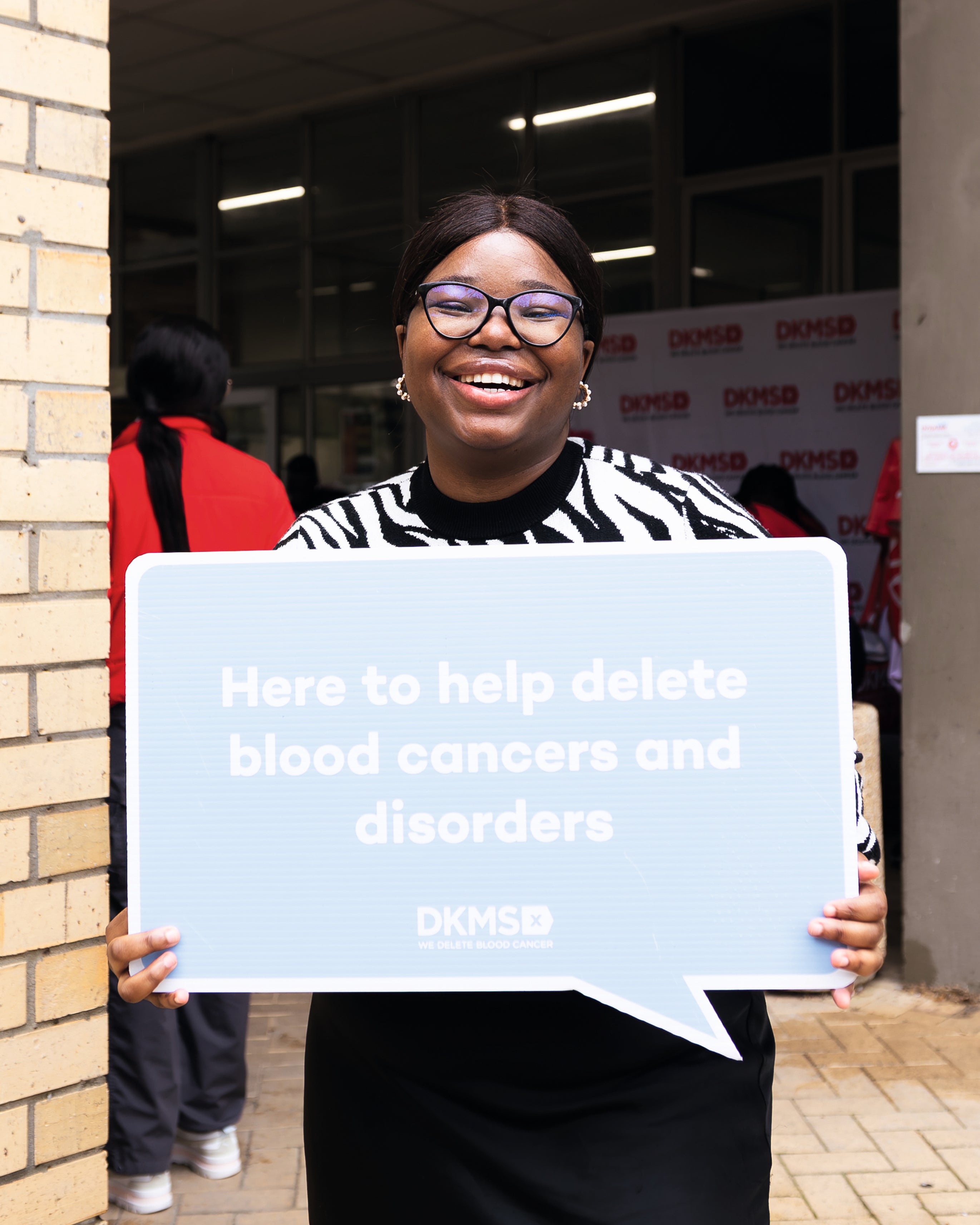 A black smiling woman wearing glasses and a black-and-white patterned sweater, holds a large light blue speech bubble sign that reads, "Here to help delete blood cancers and disorders". The DKMS logo is visible at the bottom of the sign.