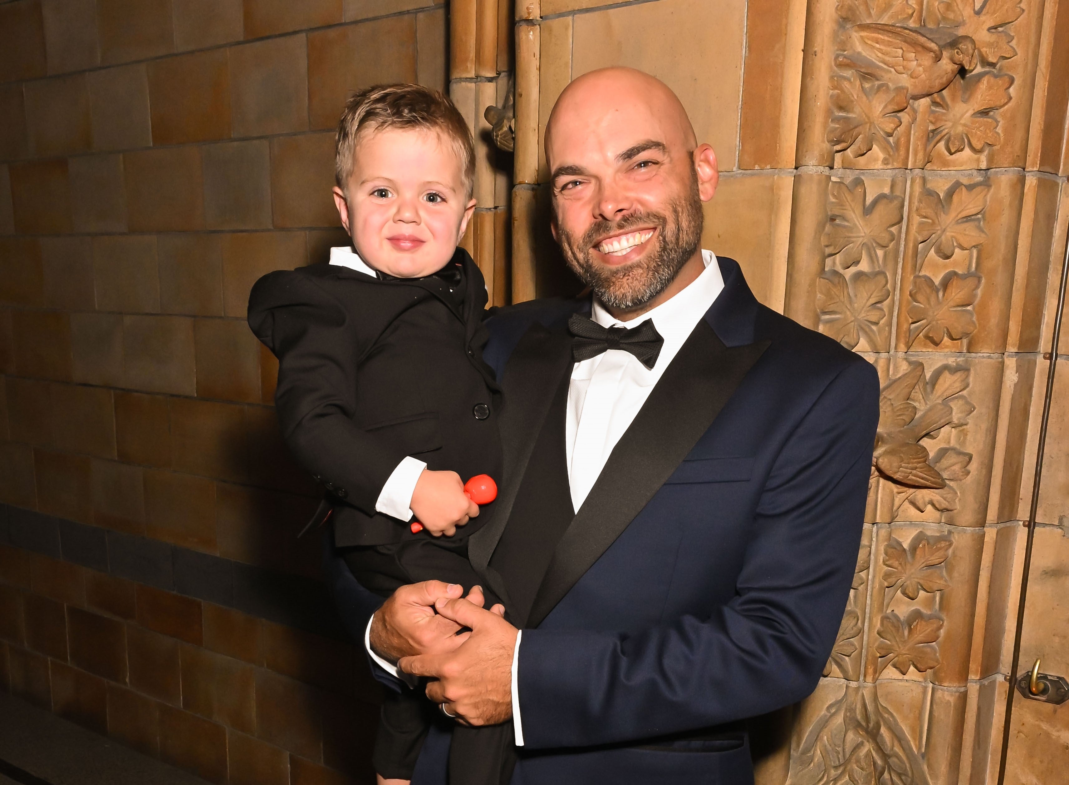 The donor Nate, wearing a navy blue tuxedo, holds the young patient Jonah Hoots. Jonah is dressed in a black suit and bow tie and is holding a small red toy. Both are posing happily for the camera in front of an ornately decorated stone wall.