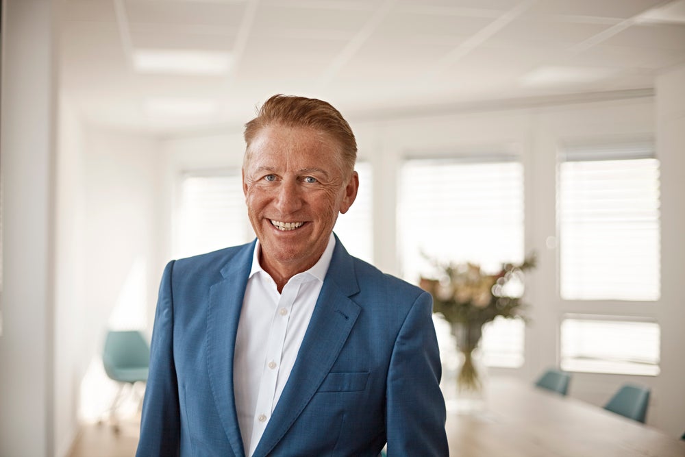 Portrait of Bernd Weinel, standing in a bright office space. He is wearing a dark blue blazer, a white shirt, and is smiling at the camera. In the background is a bouquet of flowers on a office table, blue office chairs, and windows.