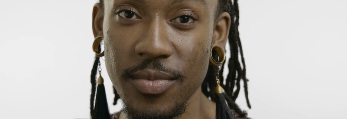 A portrait of the donor Deonté Lee. He has long, dark dreadlocks tied back in a braid, wears large hoop earrings, and a dark top. He's standing in front of a plain, light background, and looks confidently into the camera.