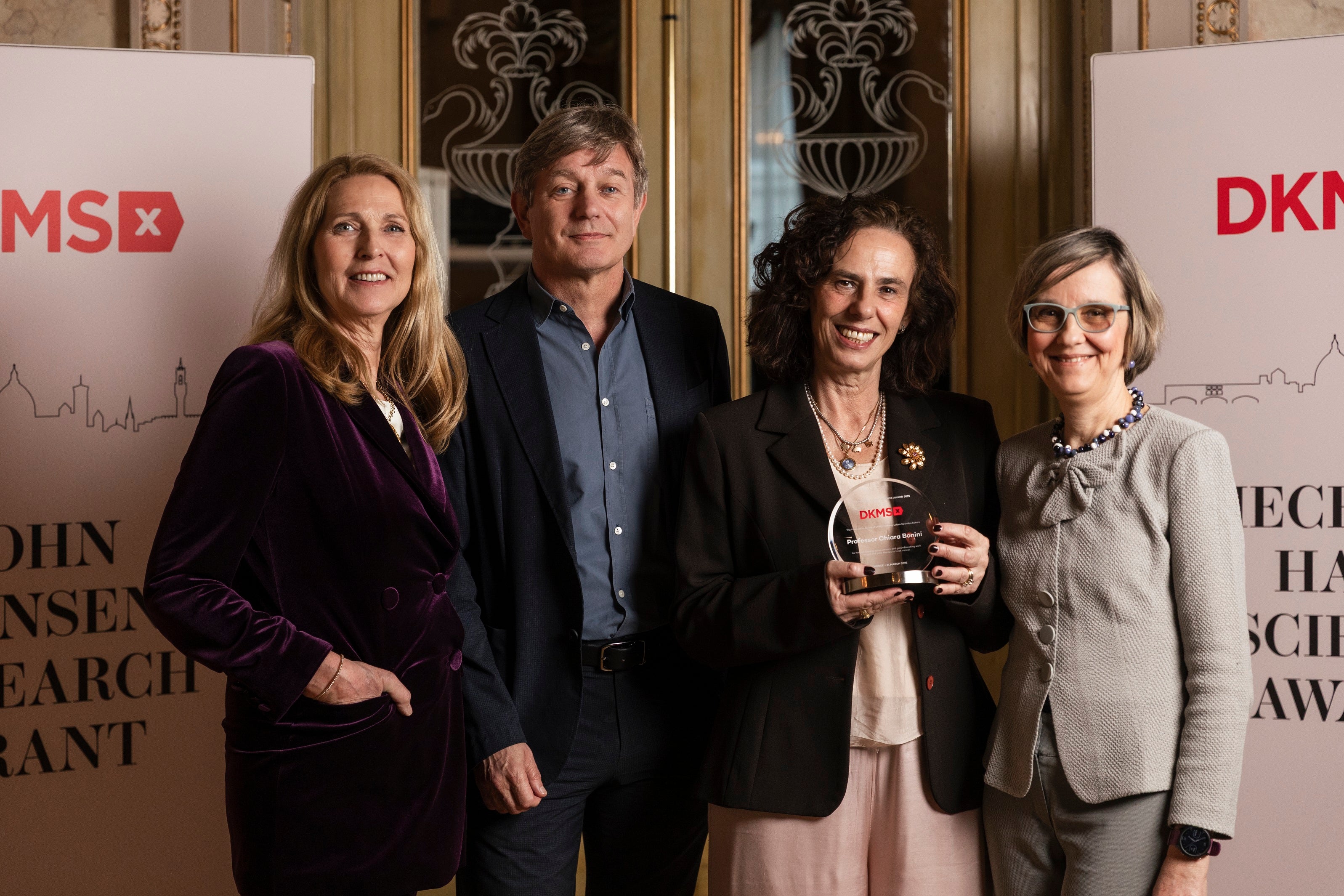 From left to right: Dr. Elke Neujahr, CEO of DKMS; Marcel van den Brink, Chair of the DKMS Medical Council; Professor Chiara Bonini, recipient of the Mechtild Harf Science Award 2025, holding the award; and Prof. Dr. Katharina Fleischhauer, standing together and smiling at the award ceremony. In the background DKMS banners and a large door are seen.