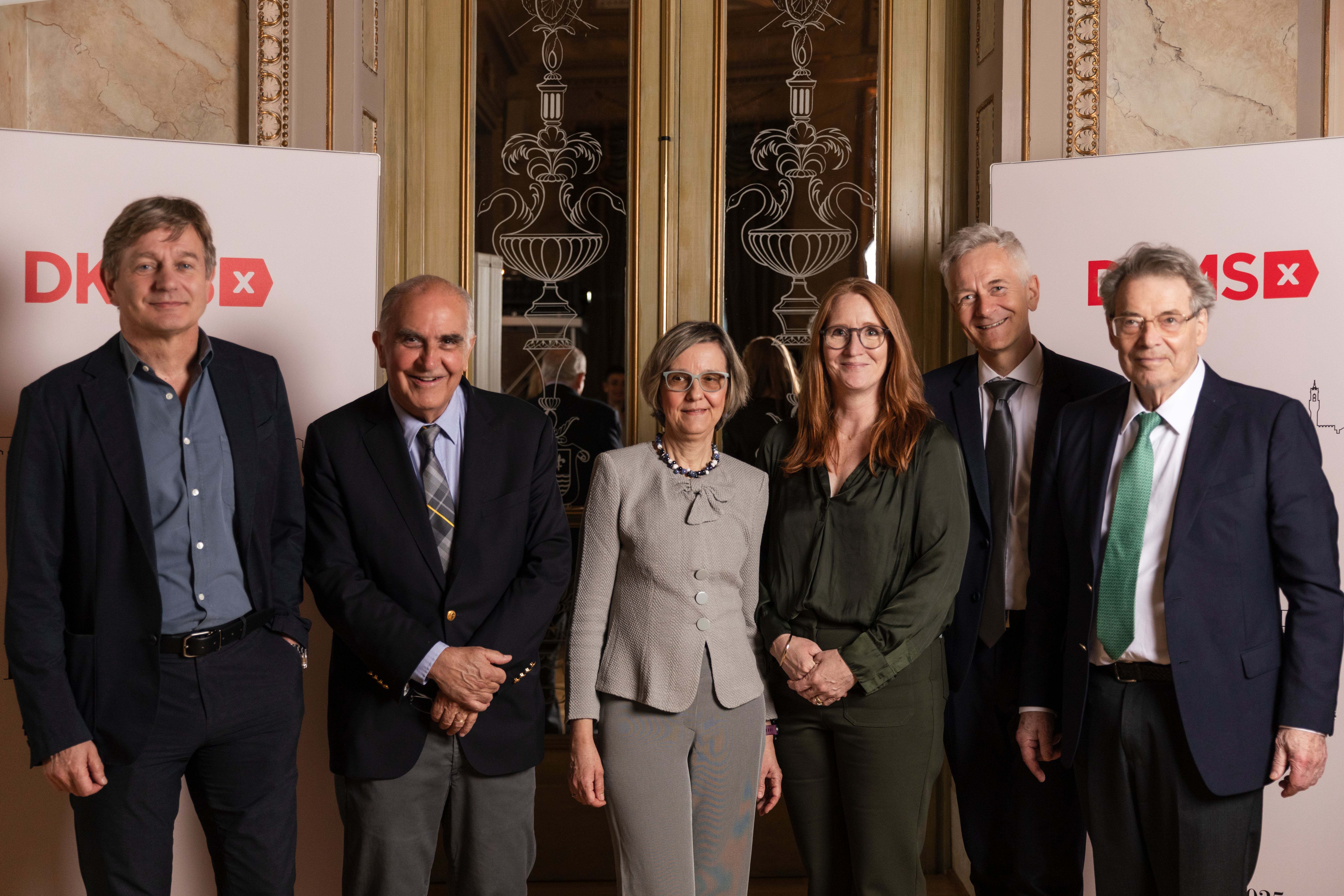 Six members of the DKMS Medical Council stand together in front of a decorative mirror background at the Mechtild Harf Science Award 2025. From the left you can see Prof. Dr. Marcel van den Brink, Dr. Marcelo Fernández-Viña, Prof. Dr. Katharina Fleischhauer, Prof. Dr. Emma Morris, Prof. Dr. Johannes Schetelig, and Prof. Dr. Dr. h.c. Dieter Hoelzer. They are dressed in business attire and smiling at the camera, with DKMS signage partially visible on both sides.