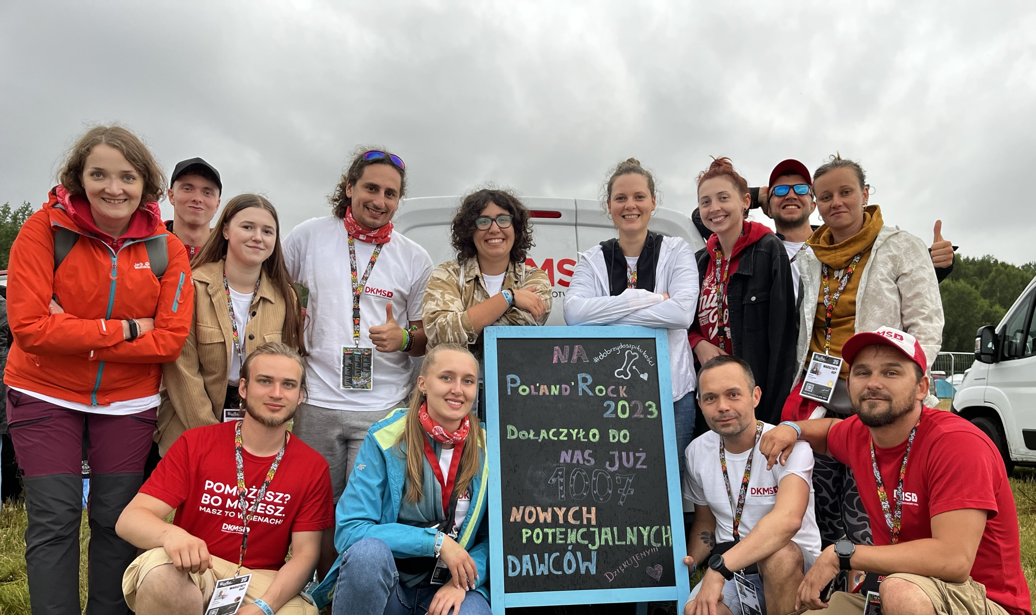 A group of thirteen enthusiastic DKMS volunteers pose together at the Poland Rock Festival 2023. They are standing and kneeling around a chalkboard that announces the number of new potential donors registered. The atmosphere is joyful and happy, with cloudy skies in the background.