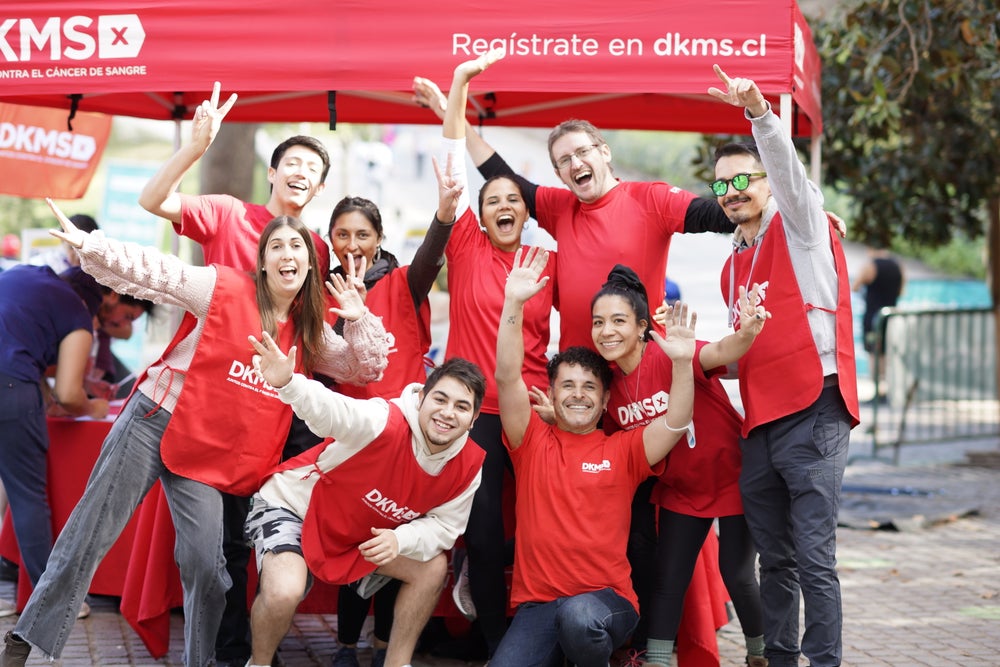 A cheerful group of nine DKMS employees poses in front of a red DKMS registration tent with their arms raised and big smiles. They are wearing red DKMS shirts and vests, and the tent reads 'Regístrate en dkms.cl,' promoting donor registration in Chile. The atmosphere is energetic and joyful.