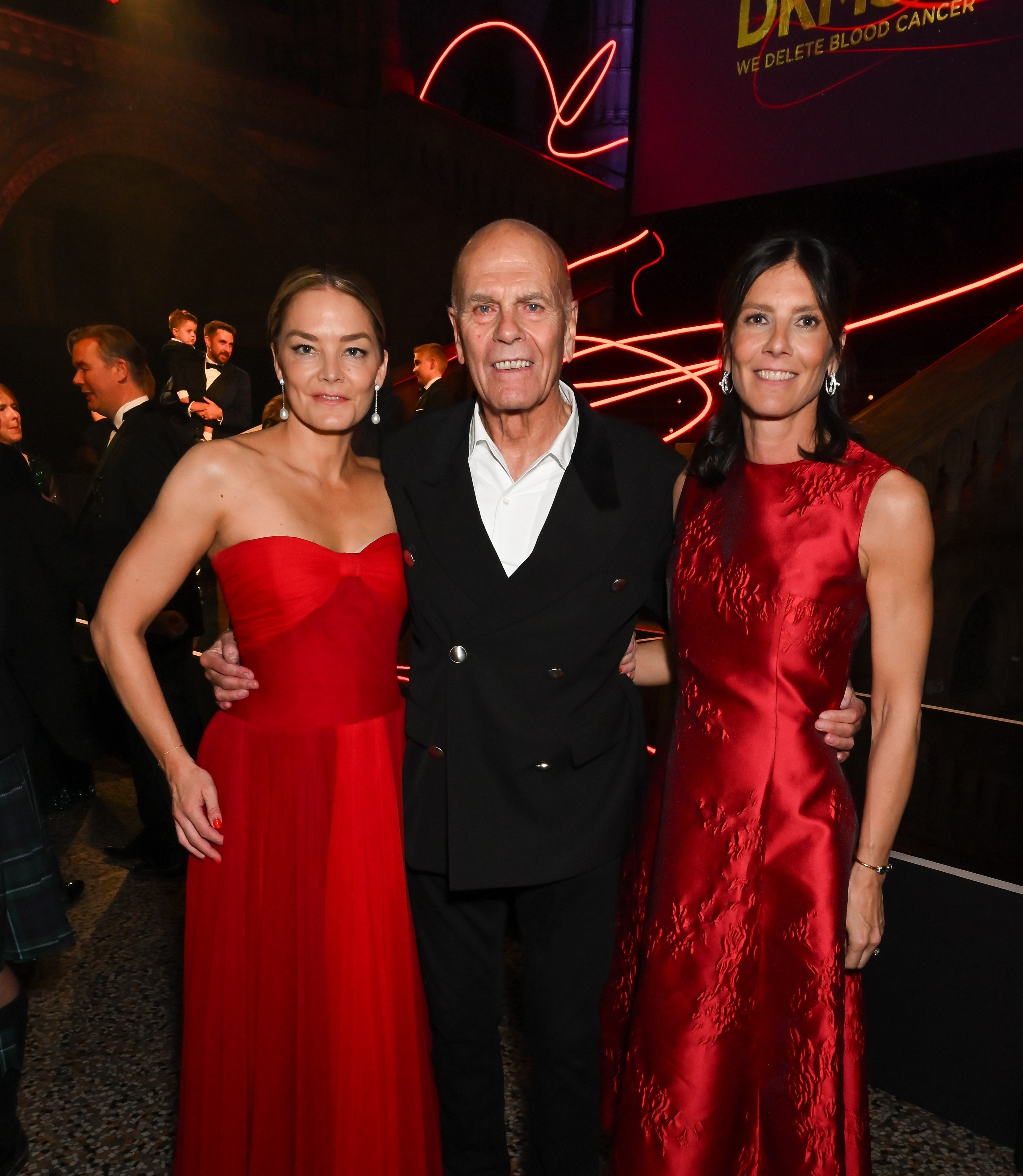 Katharina Harf, Peter Harf, and Viktoria von Wulffen pose together at the DKMS London Gala 2025. Katharina Harf and her sister Viktoria von Wulffen are dressed in elegant red evening gowns. Their father Peter Harf, who is standing in the middle, is wearing a black suit. They are smiling in front of the stage with red lightning, a part of the DKMS logo and people in the background.