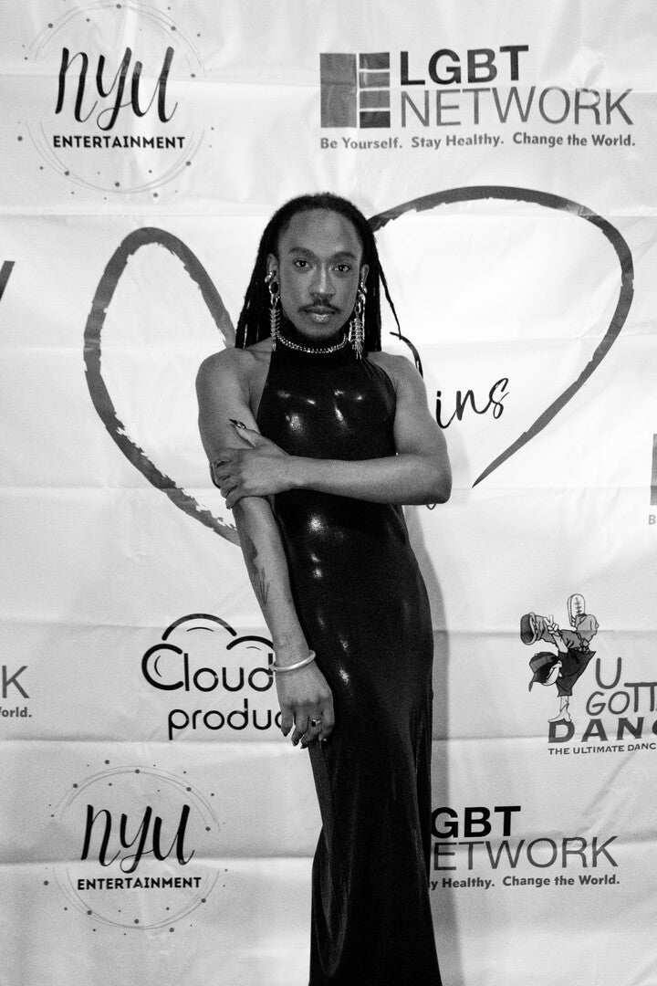 Black and white photo of the donor Deonté Lee posing confidently on a red carpet in front of a backdrop with various sponsor logos, including LGBT Network and NYU Entertainment. He's wearing a shiny black form-fitting, halter-neck gown, statement earrings, and has long dreadlocks.