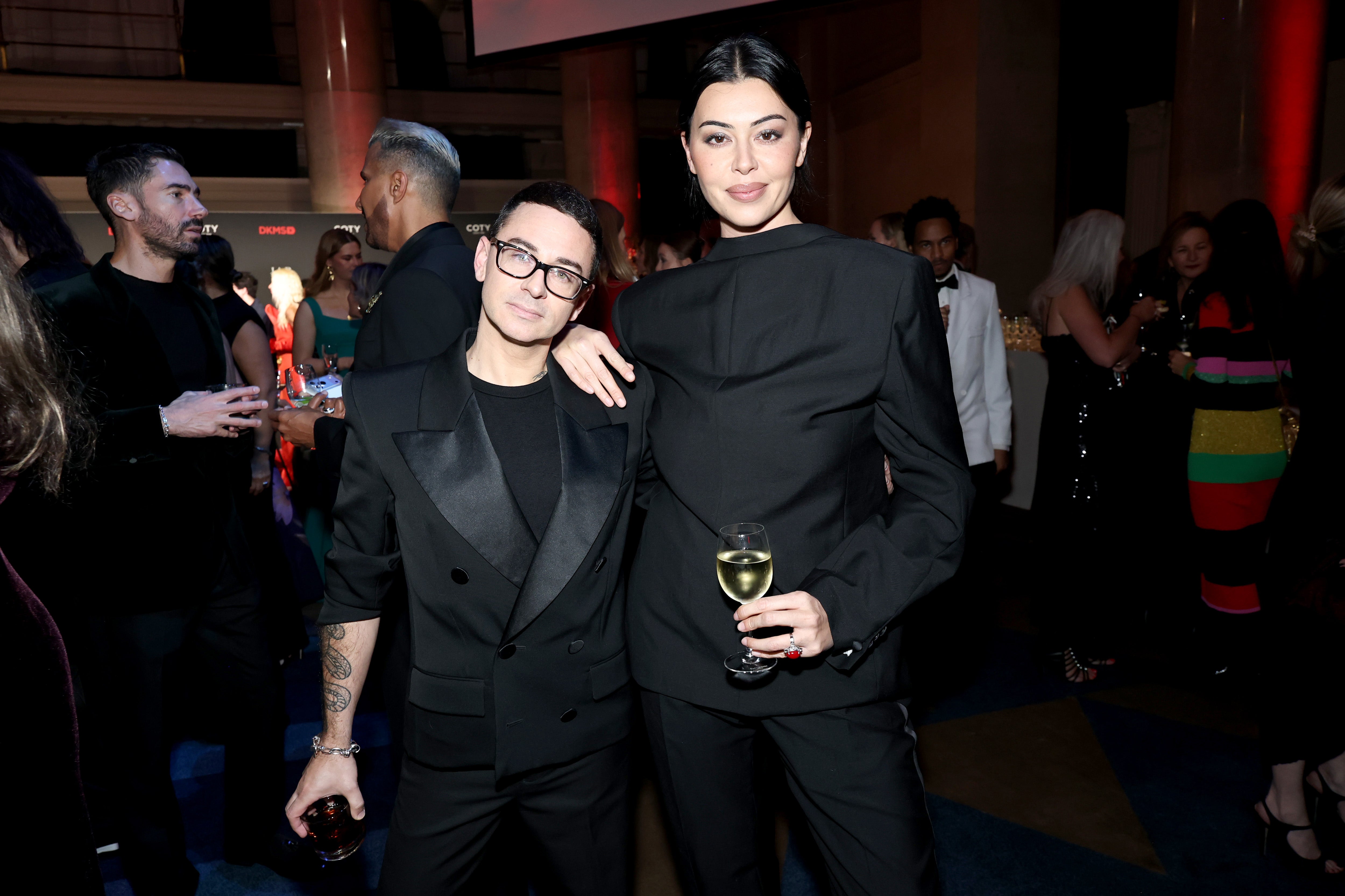 Christian Siriano and Lauren Chan, dressed in stylish black suits, pose together at the DKMS New York Gala 2025, holding drinks and smiling. Other elegantly dressed guests are visible in the background.