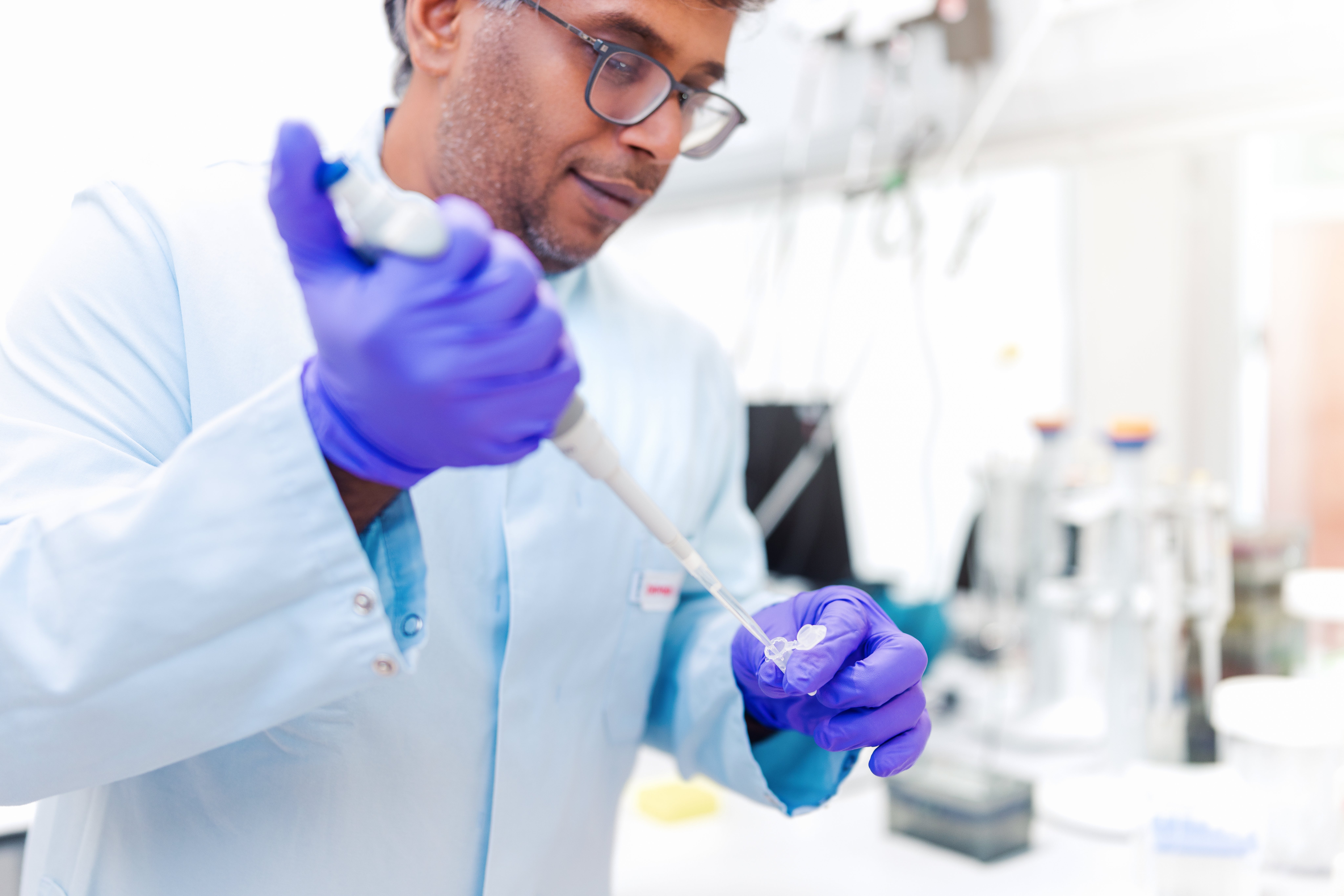 A scientist of the DKMS Life Science Lab, wearing glasses, a light blue lab coat and purple gloves, uses a pipette to transfer liquid into a small test tube in a bright laboratory.